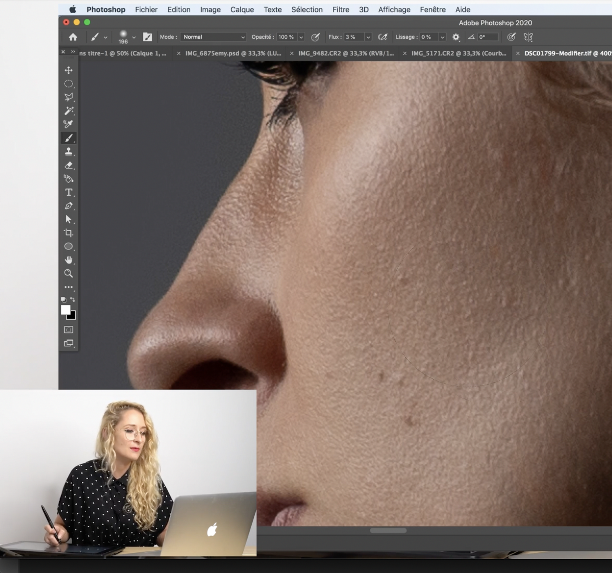 A woman with curly blonde hair and glasses sitting at a desk, working on a laptop, with a graphic design of a close-up of a person's face on a computer screen.