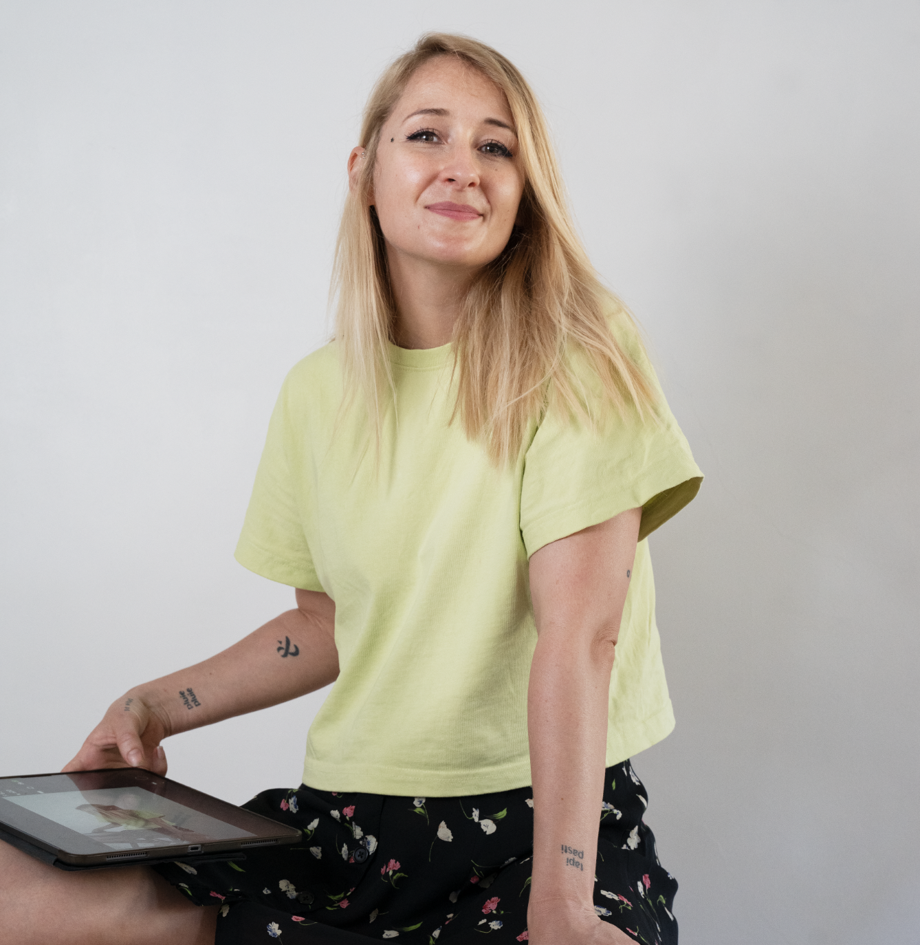 A young woman with blonde hair, wearing a yellow t-shirt and black floral pants, sitting against a plain white background, holding a tablet.