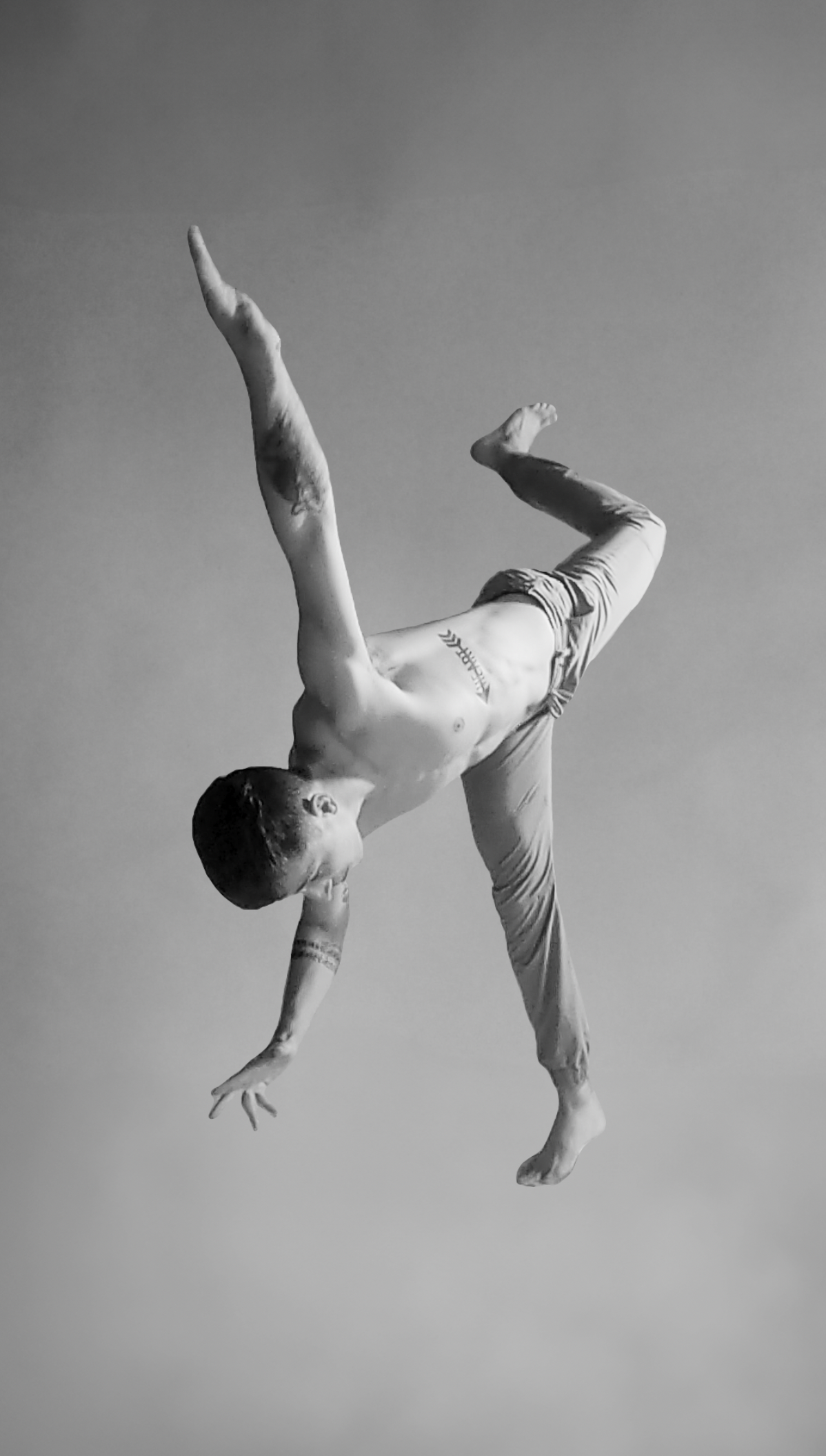 A shirtless man with tattoos is balancing on one hand in a dance or acrobatic pose against a plain background.