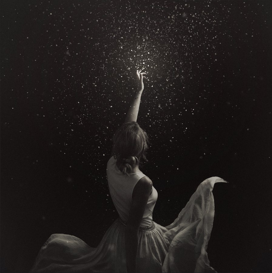A woman is sitting and reaching upward towards a shower of glittering particles against a dark background, with her back turned to the viewer, wearing a flowing dress.