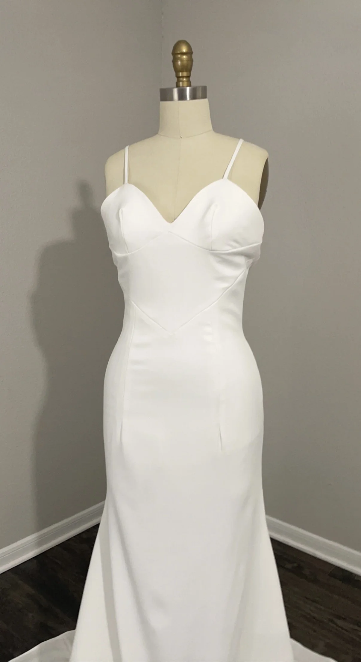 White wedding dress displayed on a mannequin in a room with beige walls and dark wood flooring.