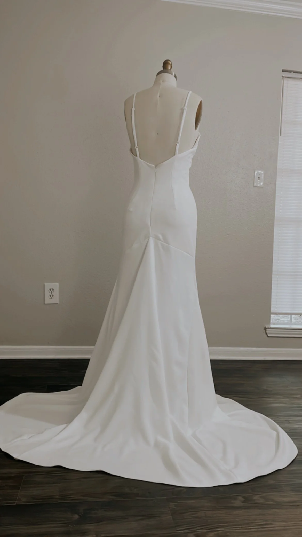 A mannequin dressed in a white wedding gown with a long train, placed in front of a plain wall in a room with wood floor and a window with blinds.