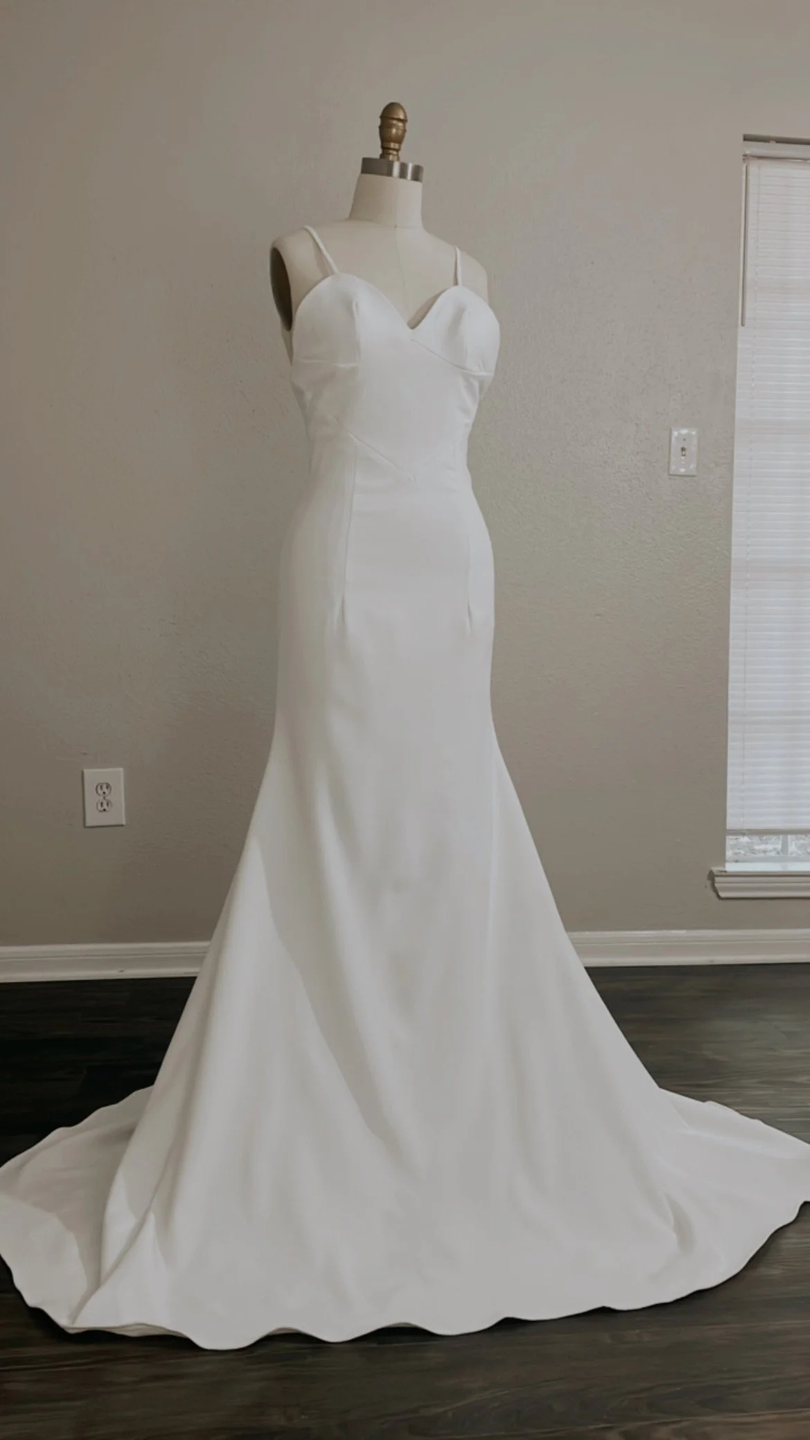 White wedding dress on a mannequin with a long train in a room with beige walls and hardwood floors.