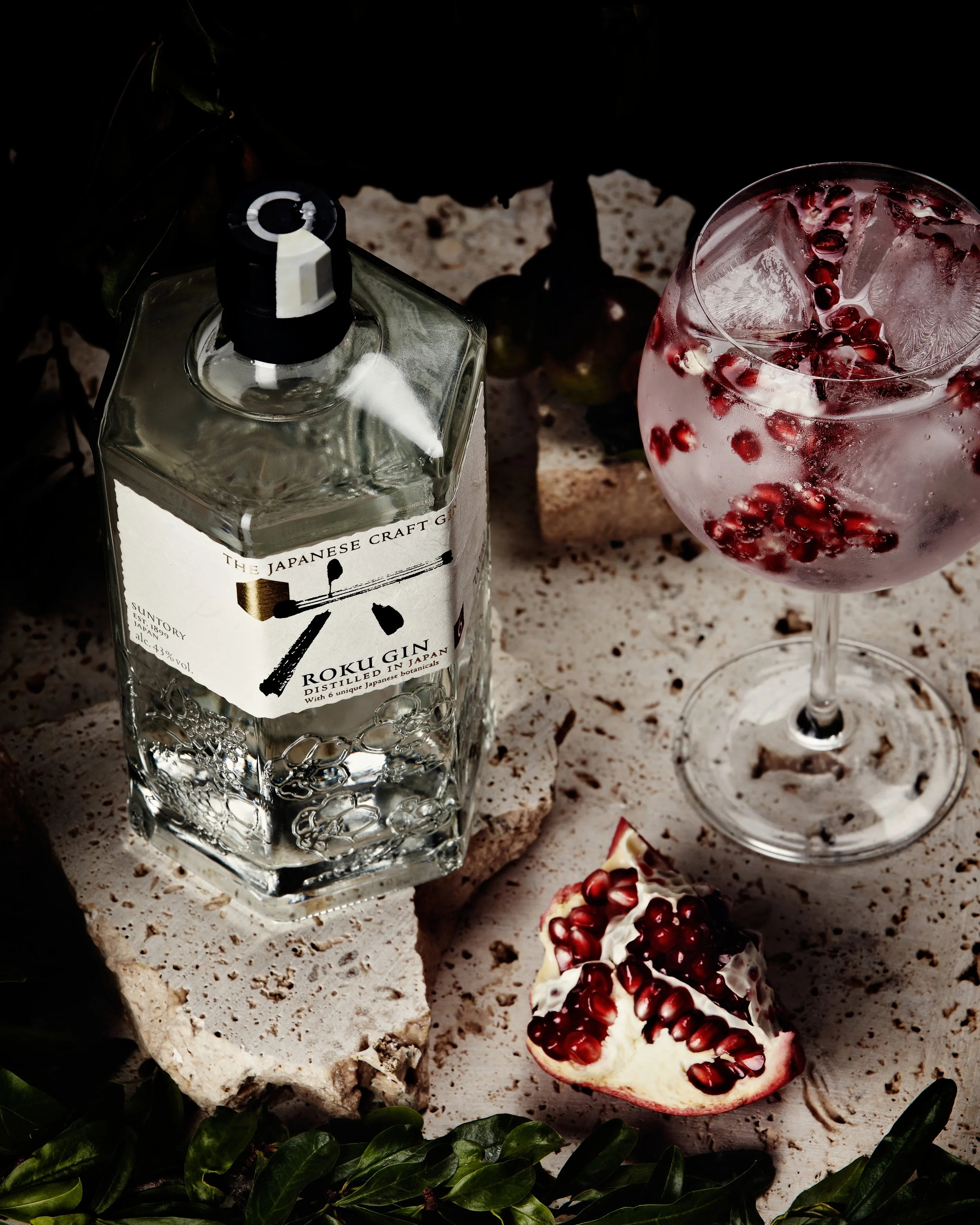 Image Description: This image contains a gin spritz filled with pomegranate, next to a bottle of Roku Japanese gin and decorated with leaves and white stone