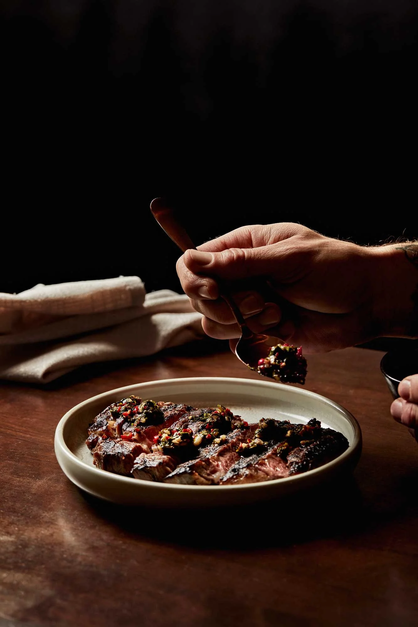 Image Description: This image contain a chargrilled steak that has been sliced and plated with a chef drizzling chimichurri over the top with a napkin in the background.