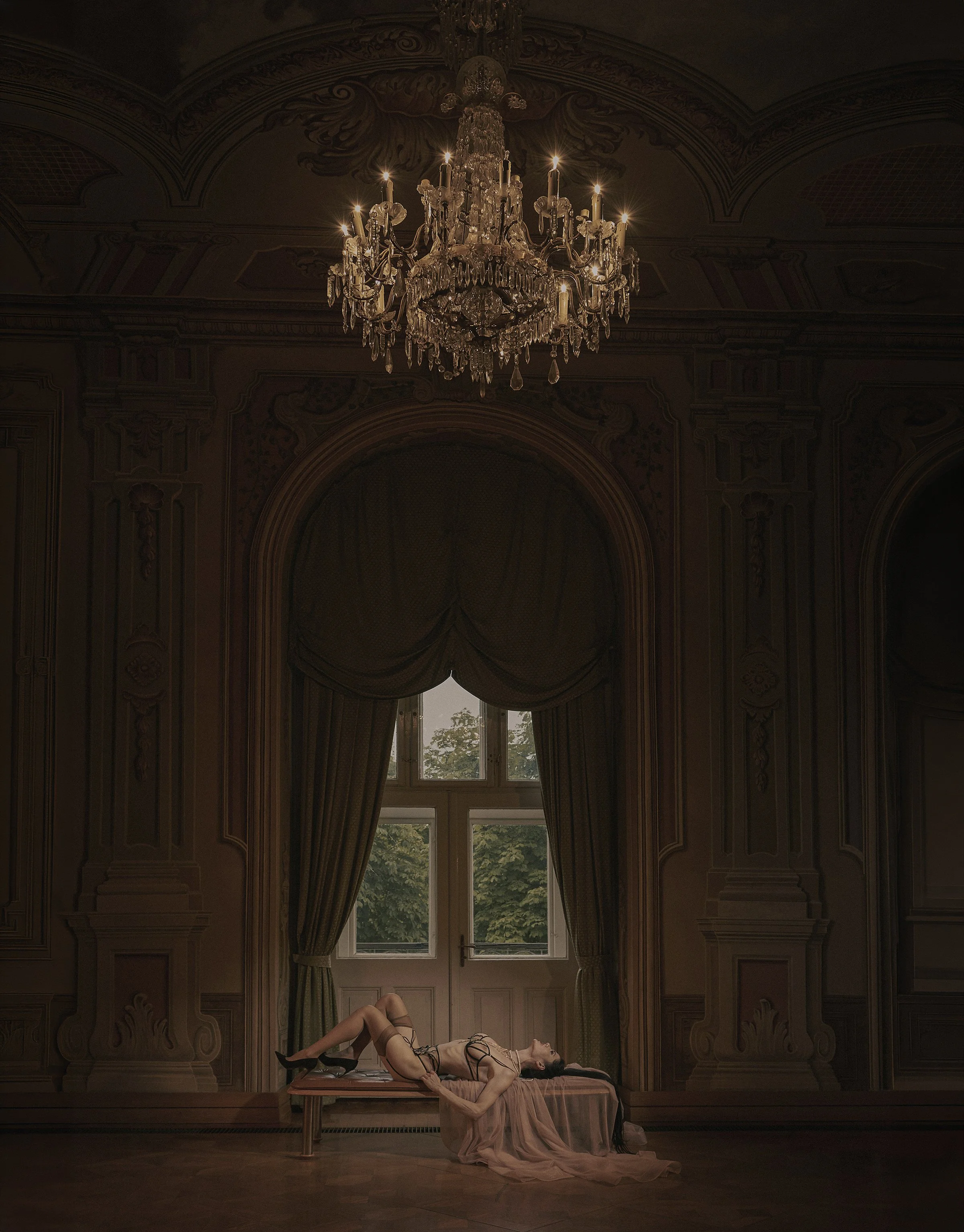 Fine-art boudoir portrait of a woman reclining beneath a crystal chandelier in an ornate historic interior.