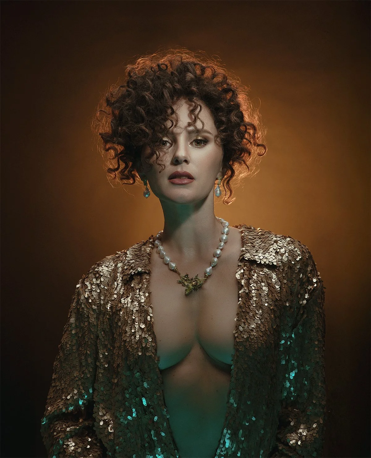 Boudoir portrait of a woman with curly hair wearing a sequined gold jacket and pearls, captured in warm vintage-style lighting
