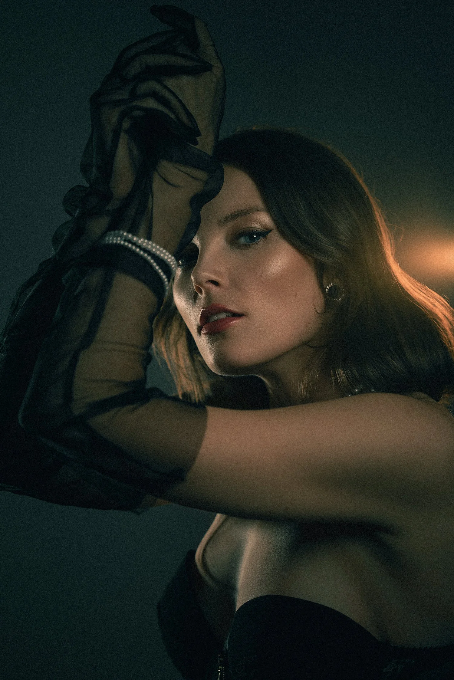 Editorial boudoir portrait featuring a woman in black lingerie and sheer gloves, dramatic low-light setting and refined, intimate styling.