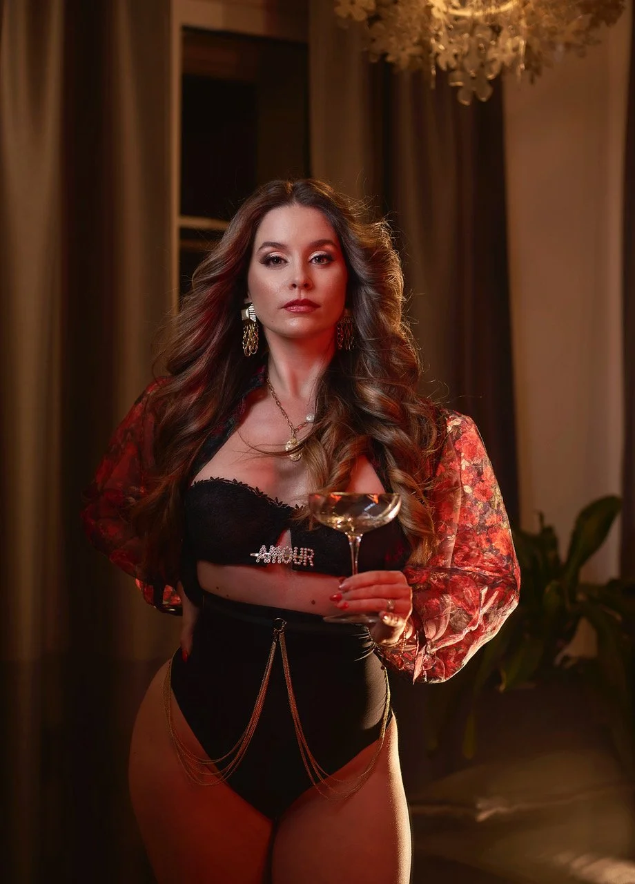 Woman with long hair holding a champagne glass, wearing a black outfit and floral sheer sleeves, standing indoors.