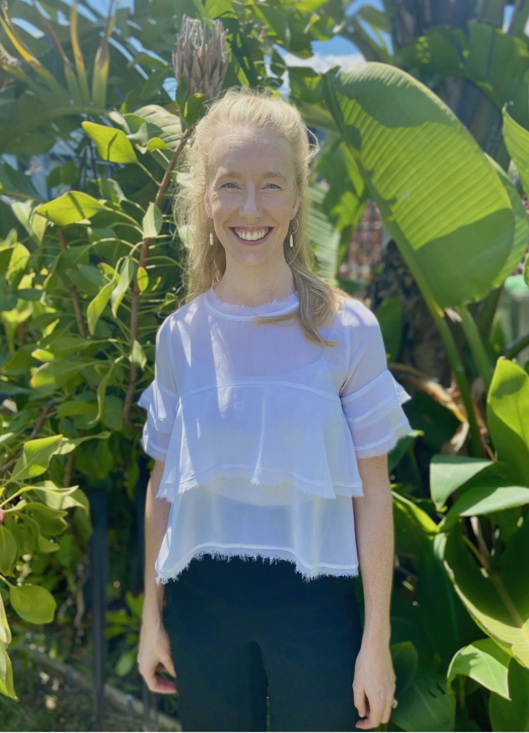 A woman with blonde hair smiling outdoors in front of lush green tropical plants, wearing a white sheer blouse and hoop earrings.