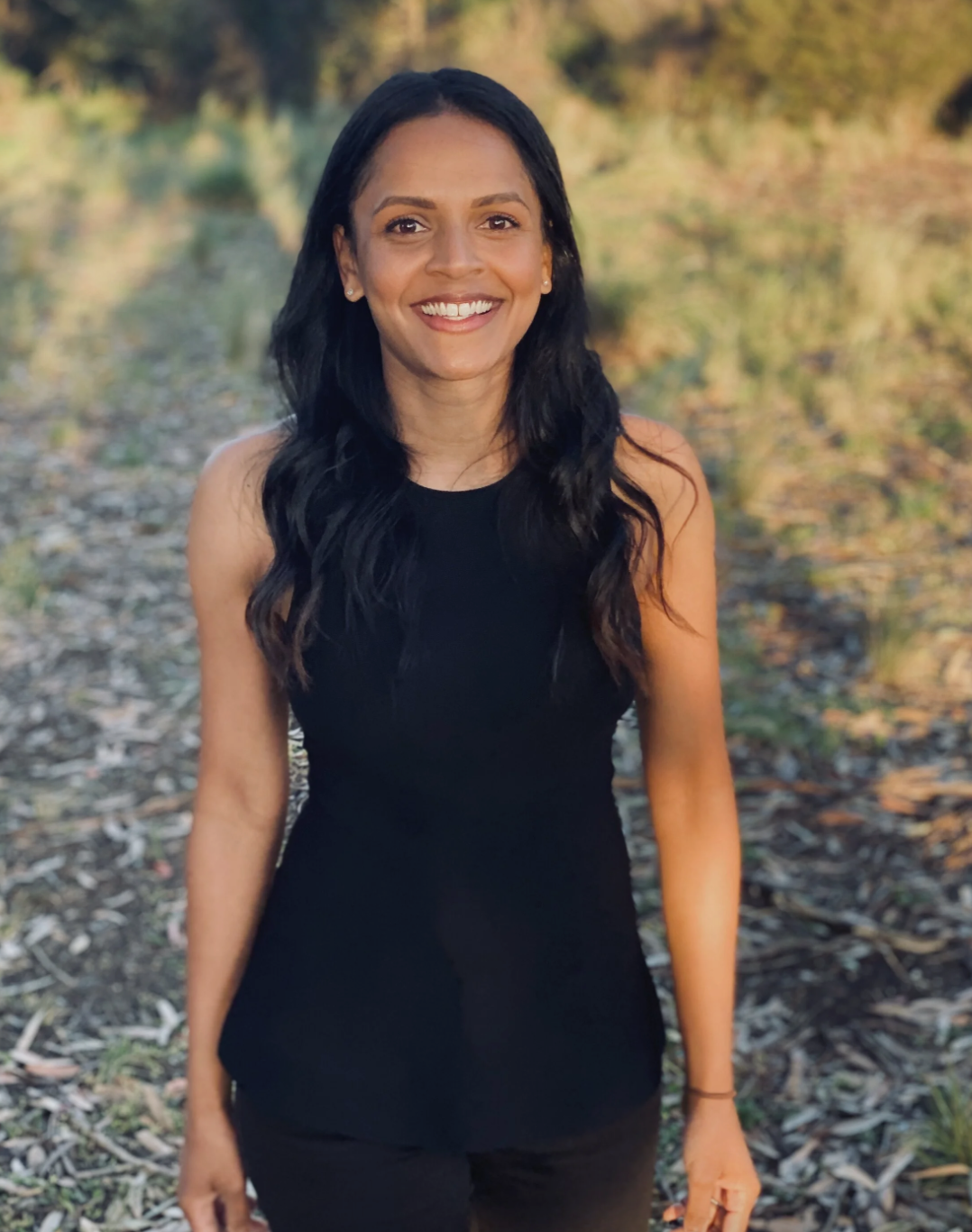 A woman smiling outdoors in a natural setting with trees and fallen leaves.