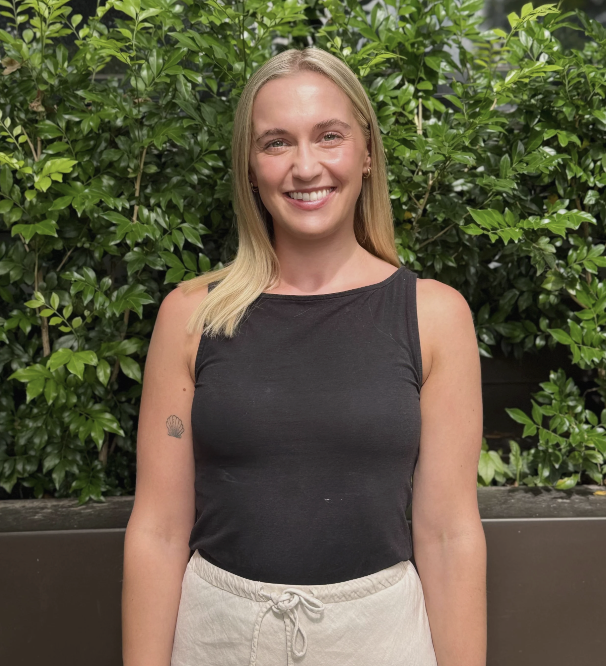 A young woman with blonde hair, smiling, wearing a black sleeveless top and beige drawstring shorts, standing in front of green leafy plants.