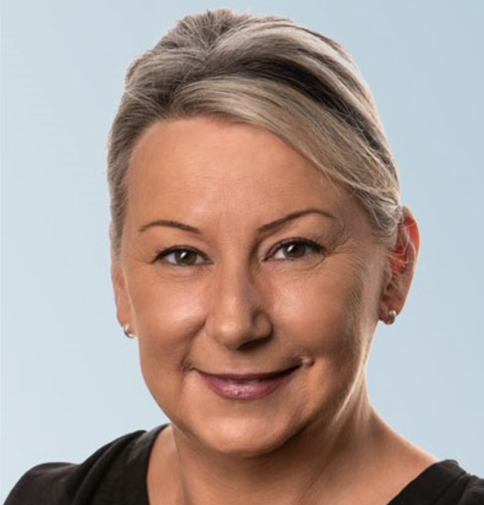 Close-up portrait of a middle-aged woman with short, gray hair, smiling, wearing small earrings, and a black top against a light blue background.