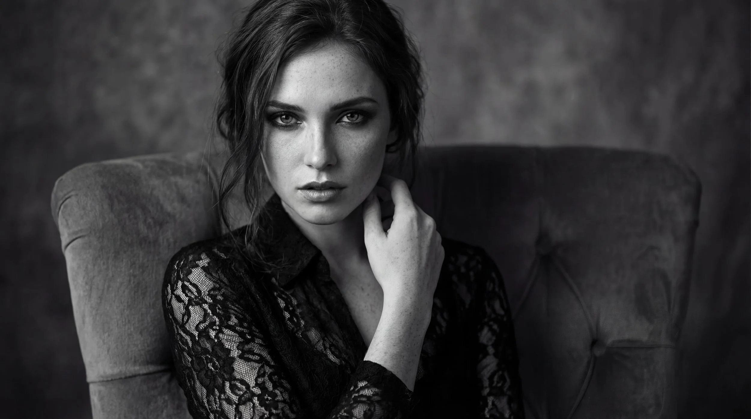 Schwarz-weiß-Foto einer jungen Frau mit dunklen, lockigen Haaren, die auf einem bequemen Sessel sitzt, trägt ein schwarzes Spitzenoberteil, schaut ernst und direkt in die Kamera, Hintergrund ist unscharf.