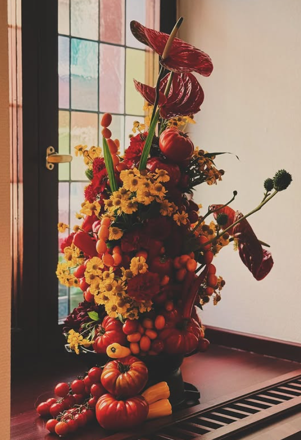 A floral arrangement with various vegetables and flowers, including tomatoes, pumpkins, calla lilies, yellow daisies, and anthuriums, placed on a wooden surface near a window.