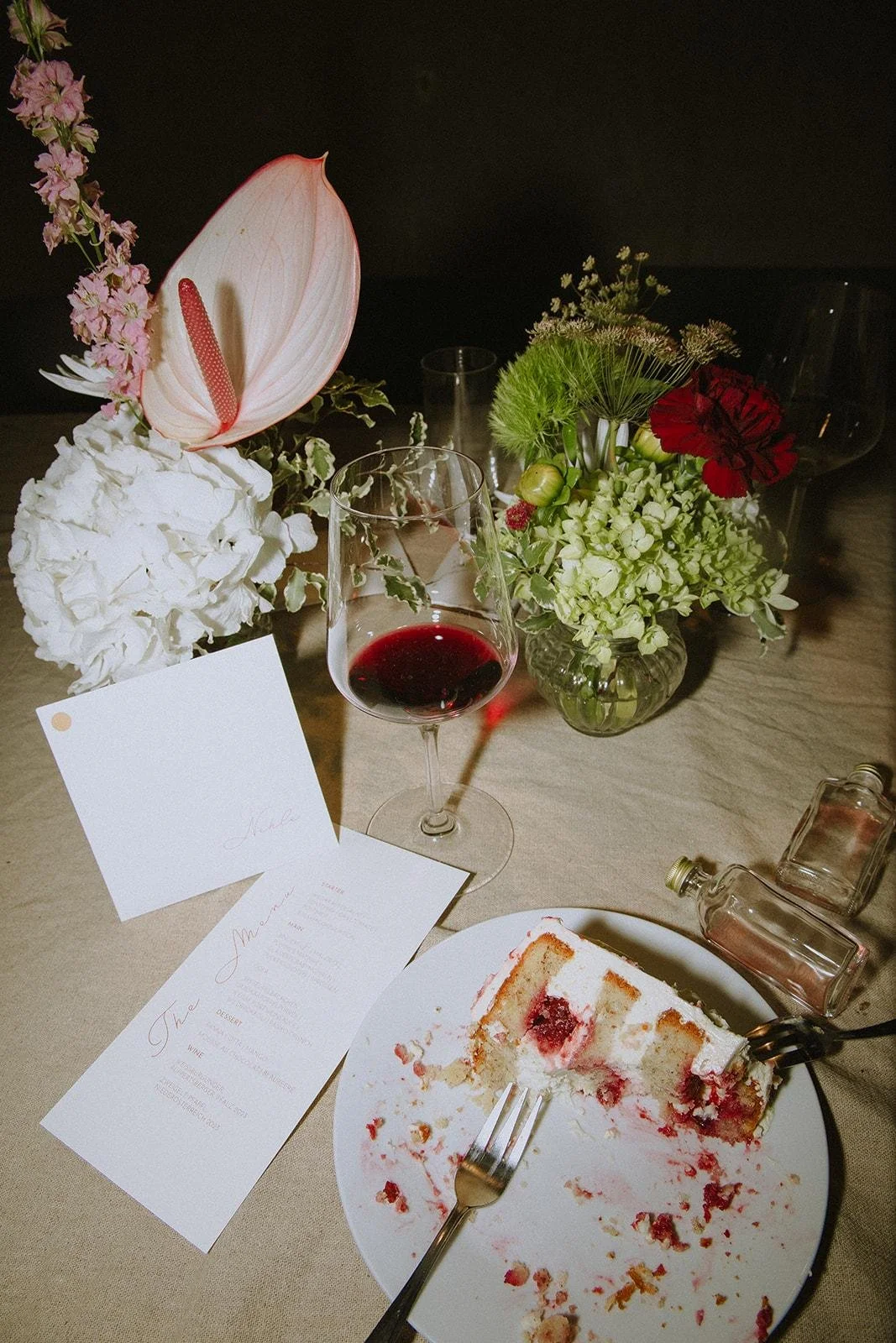 A partially eaten slice of cake on a white plate, a glass of red wine, floral centerpieces, and a menu card on a beige tablecloth.