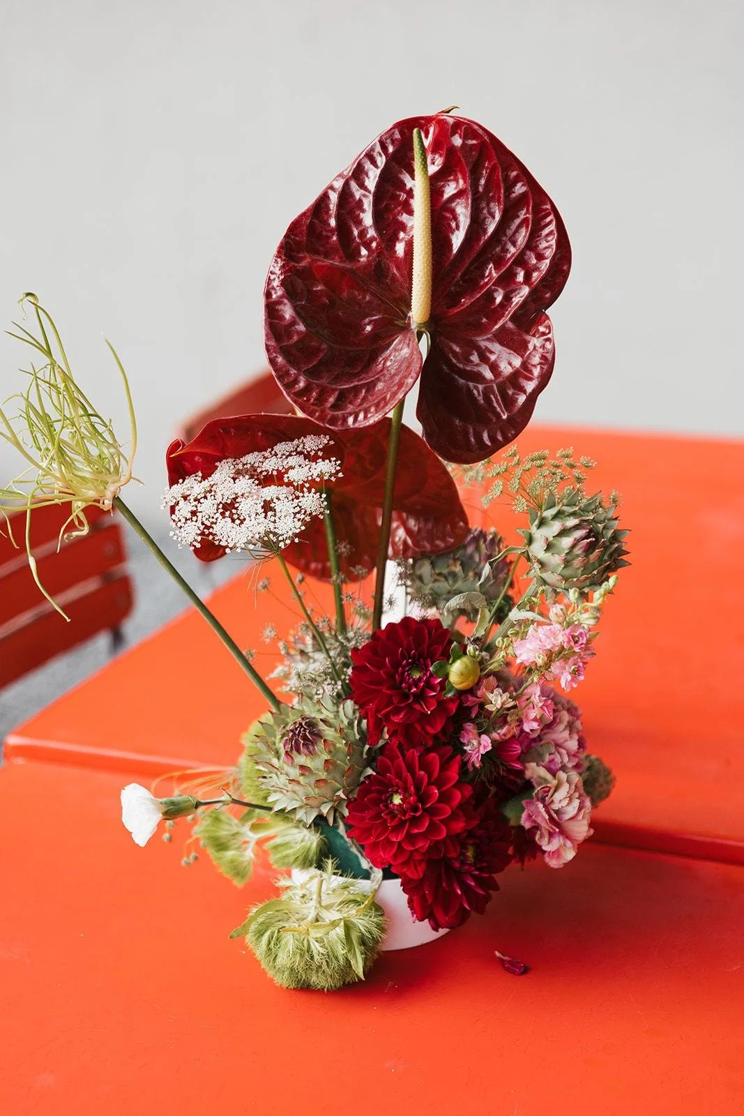 A floral arrangement with red, pink, and green flowers and plants, placed on an orange table.