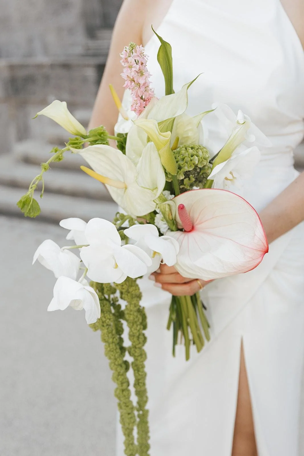 A person in a white dress holding a bouquet of various white and pink flowers, including anthuriums, calla lilies, and orchids, with a blurred stone background.