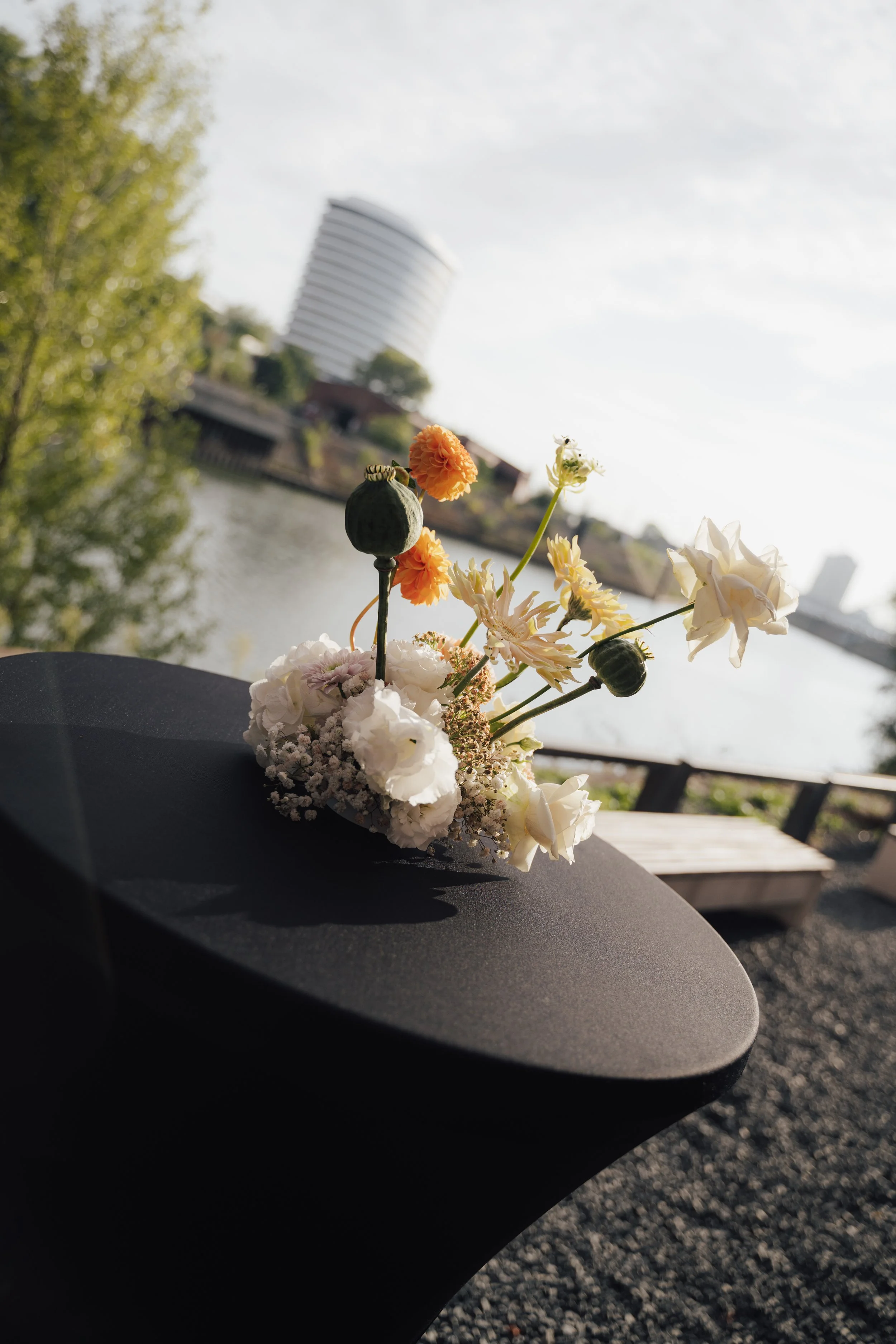 A small floral arrangement with white, orange, and pale yellow flowers placed on a black table outdoors near a body of water, with trees and a modern white high-rise building in the background under a partly cloudy sky.