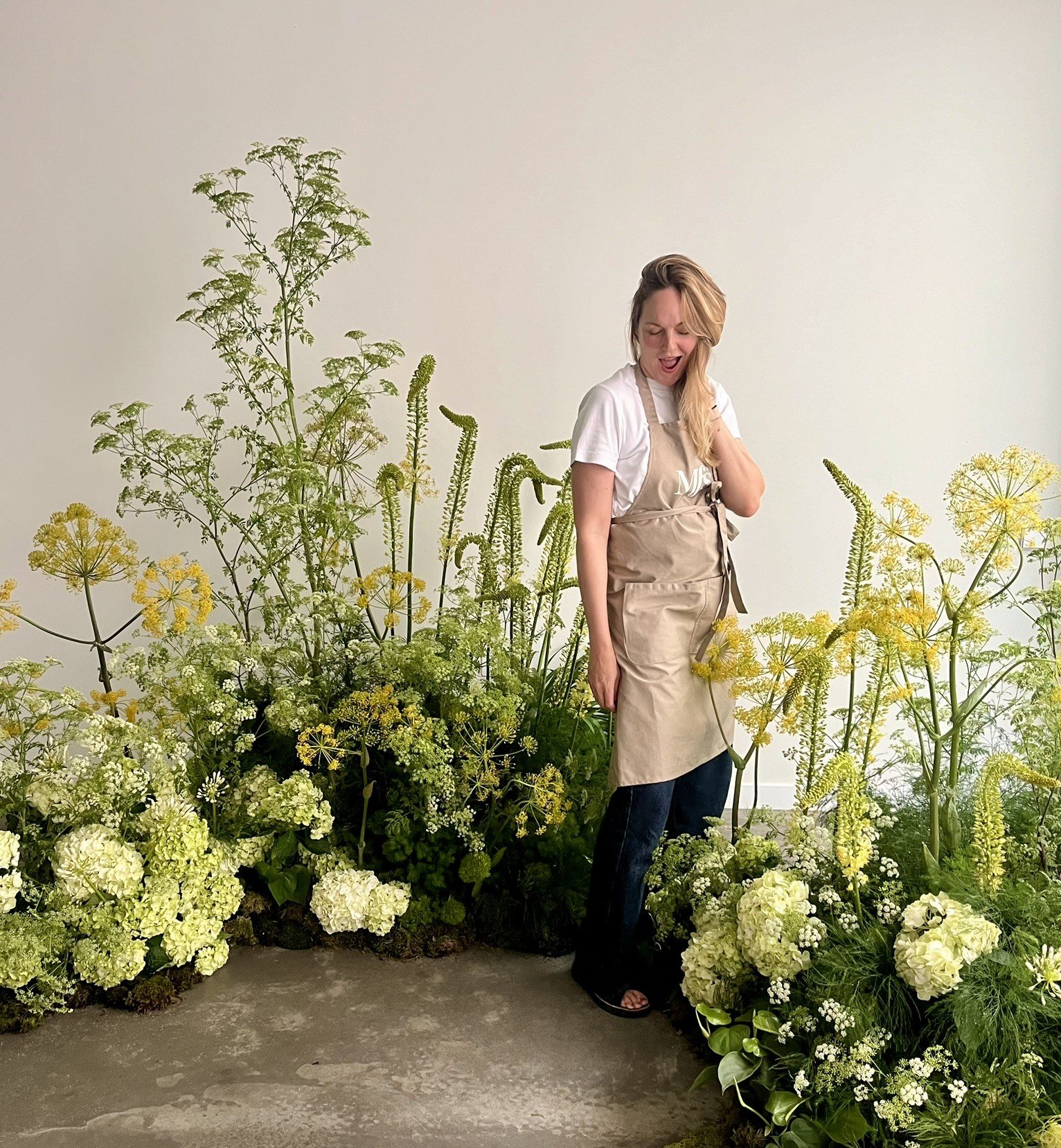 Woman in a white t-shirt and beige apron standing among green and white flowering plants indoors, looking down with a surprised expression.