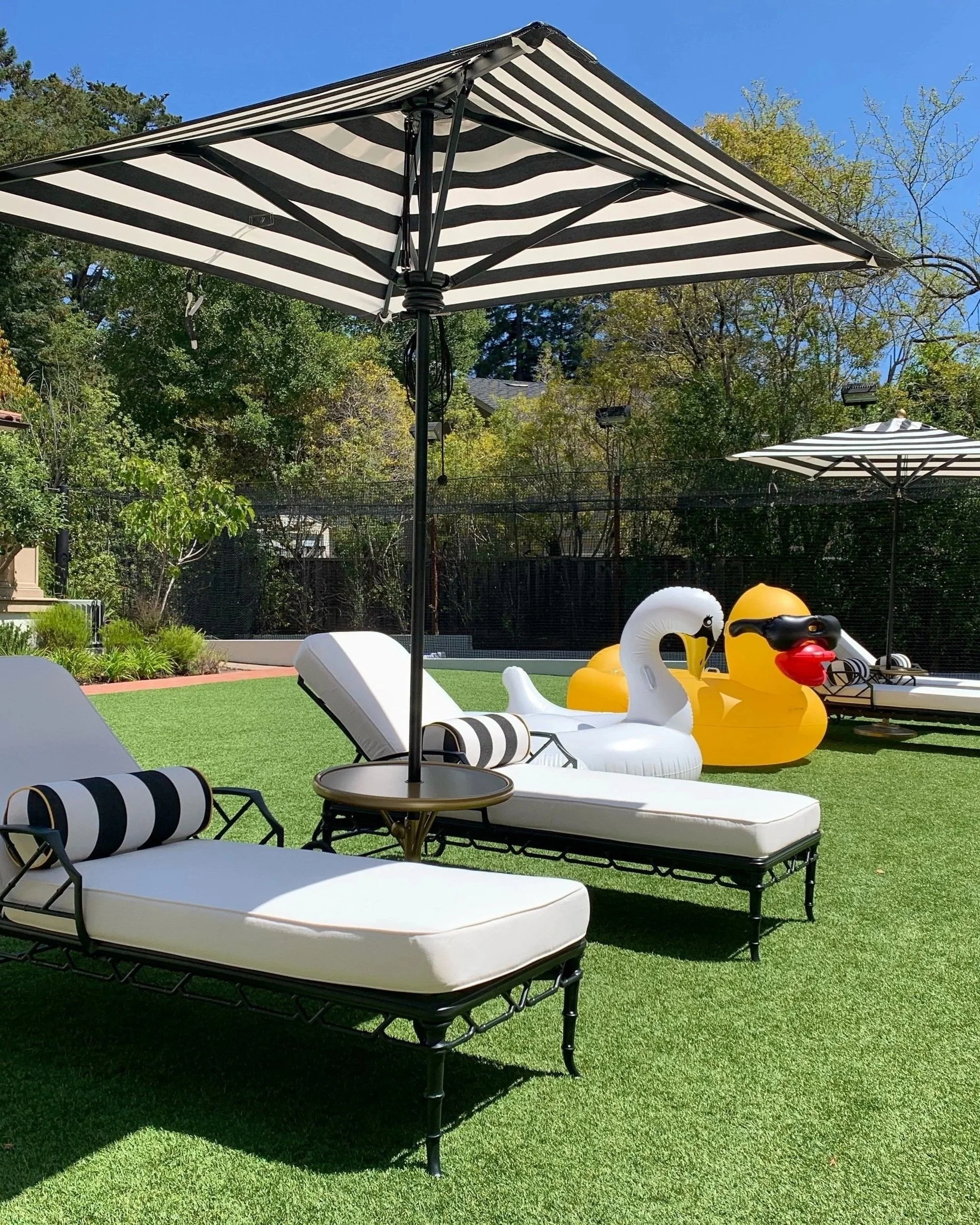 Outdoor chaise lounges in black bamboo with white cushions and striped accent pillows. A large yellow rubber duckie pool float and a white swan pool float sit ready for play.