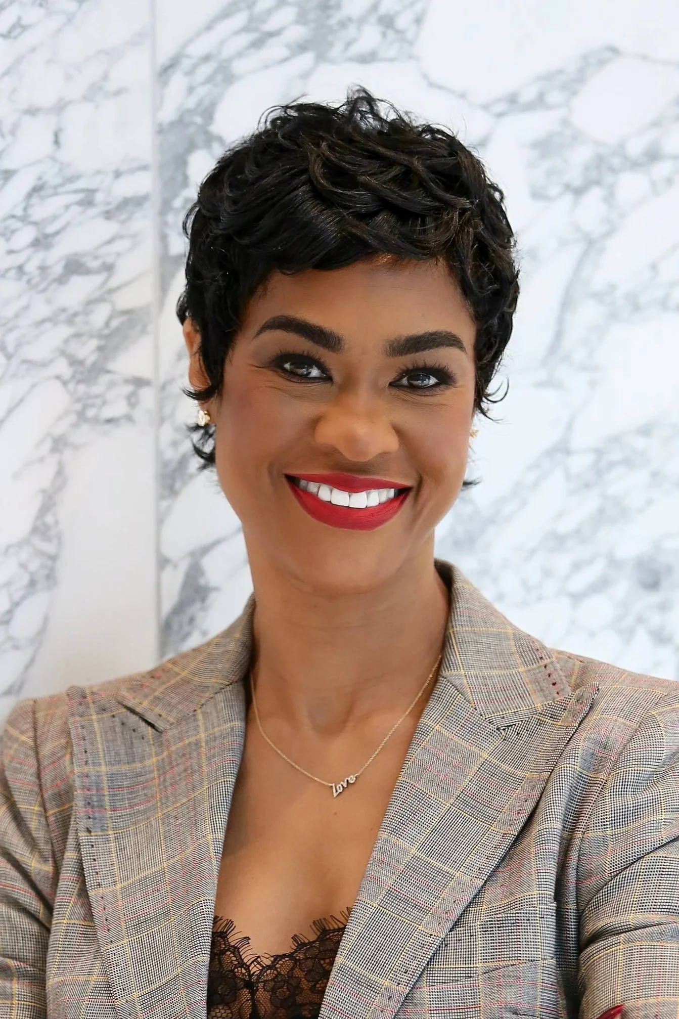 A woman with short curly black hair and red lipstick is smiling at the camera. She wears a black blazer with patterned fabric and a delicate necklace.