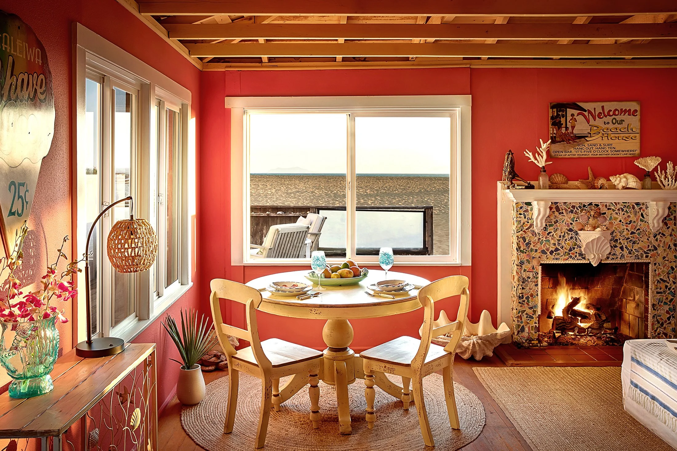 Bright living room with red walls, yellow ceiling beams, and beach-themed decor, including surfboards and tropical artwork. There is a white sofa with colorful pillows, a coffee table with magazines, and a yellow dining table with chairs. Large windows overlook a patio area with a beach-themed surfboard on the wall.
