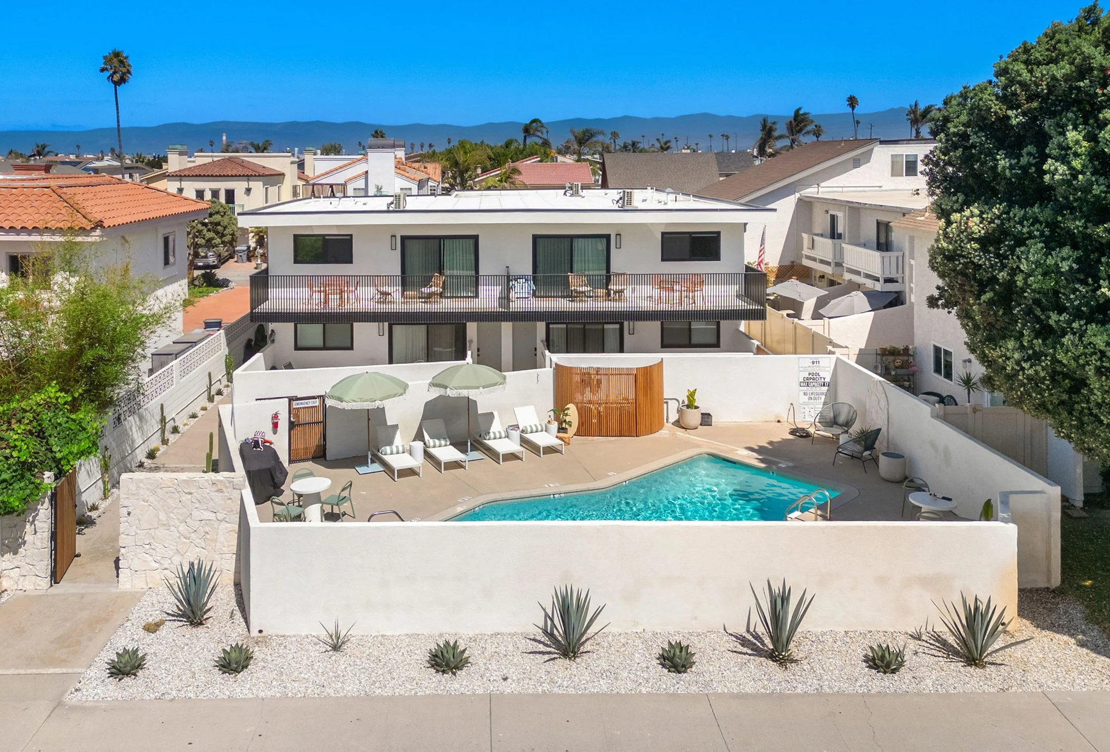 Open-concept living and dining area with beige and natural wood furniture, a large window with curtains, a wall-mounted TV, and a small kitchen in the background, decorated with plants and neutral-toned decor.
