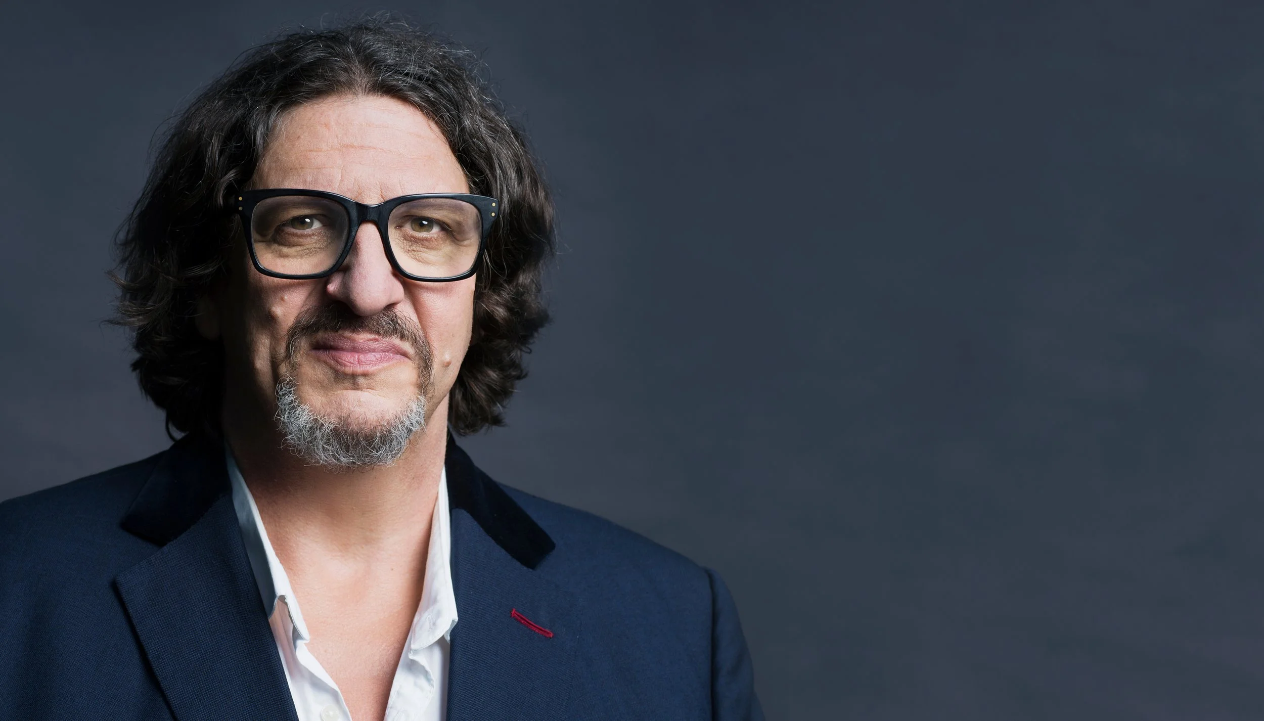 A middle-aged man with shoulder-length wavy hair, wearing large black glasses, a navy blazer with a red stitch on the lapel, and a white dress shirt, standing against a dark gray background.