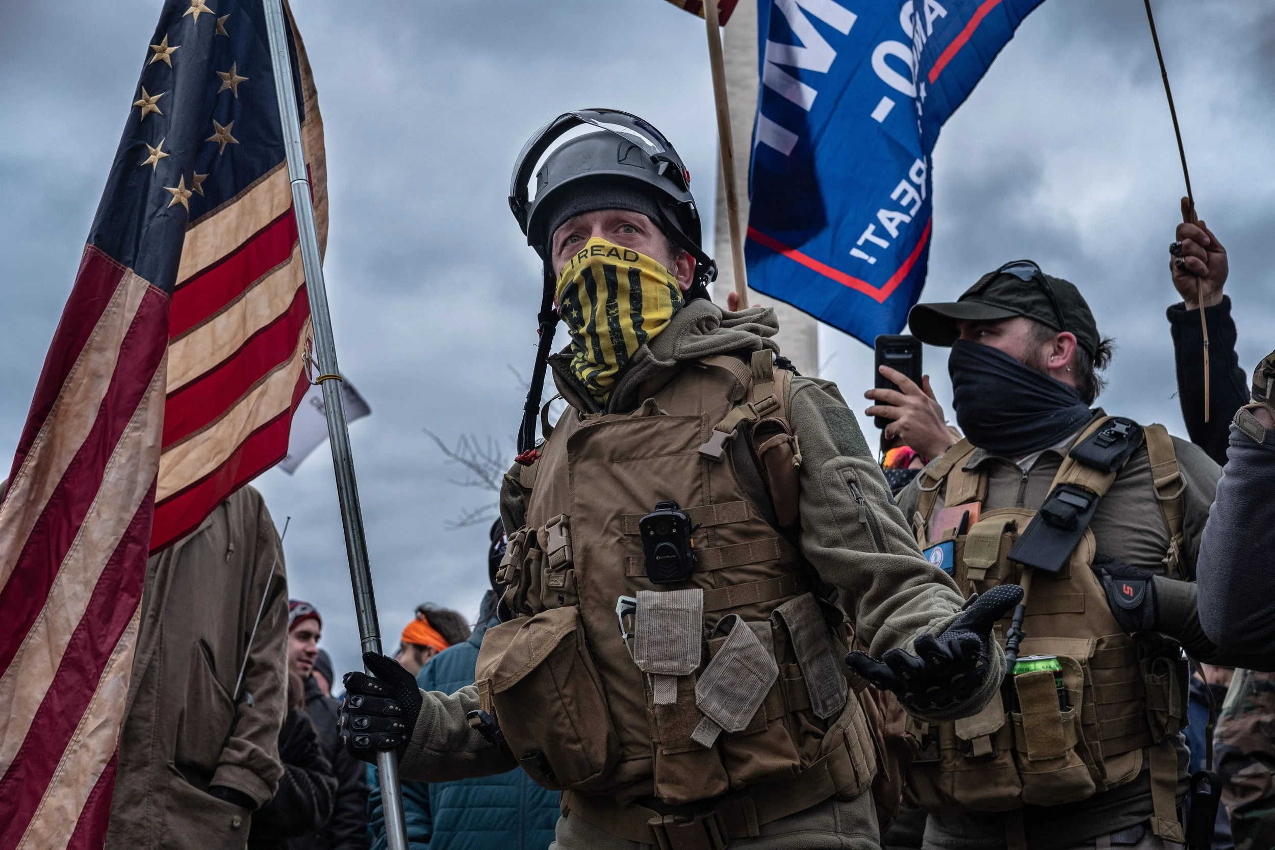 Group of people, some wearing tactical gear, holding flags and banners in an outdoor setting with a cloudy sky.