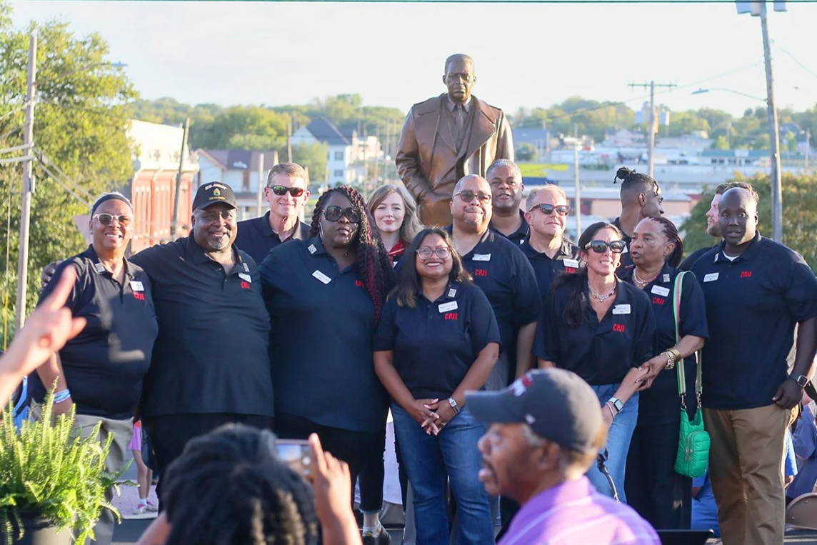 Group of diverse people wearing black shirts with red and white name tags, standing outdoors in front of a statue and small town buildings, during daylight.