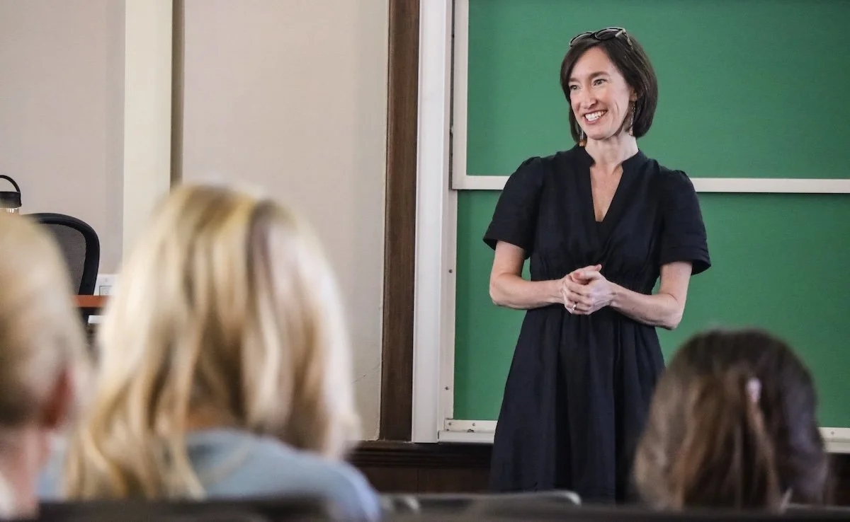 A woman with short brown hair, wearing a black dress, stands in front of a green chalkboard in a classroom, smiling and speaking to an audience of students.