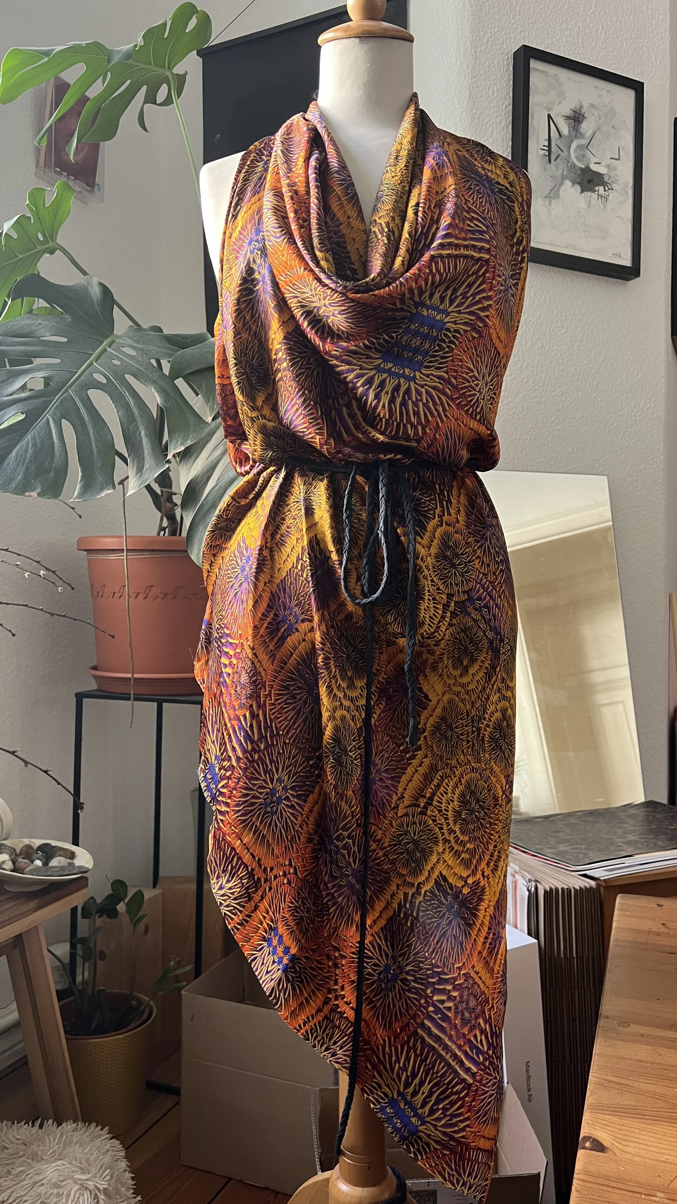 A mannequin dressed with a colorful, mushroom inspired patterned fabric in gold and purple hues,  draped around the shoulders and waist. This almost psychedelic print is a new take on plaid but is elevated with gills and a graphic touch.