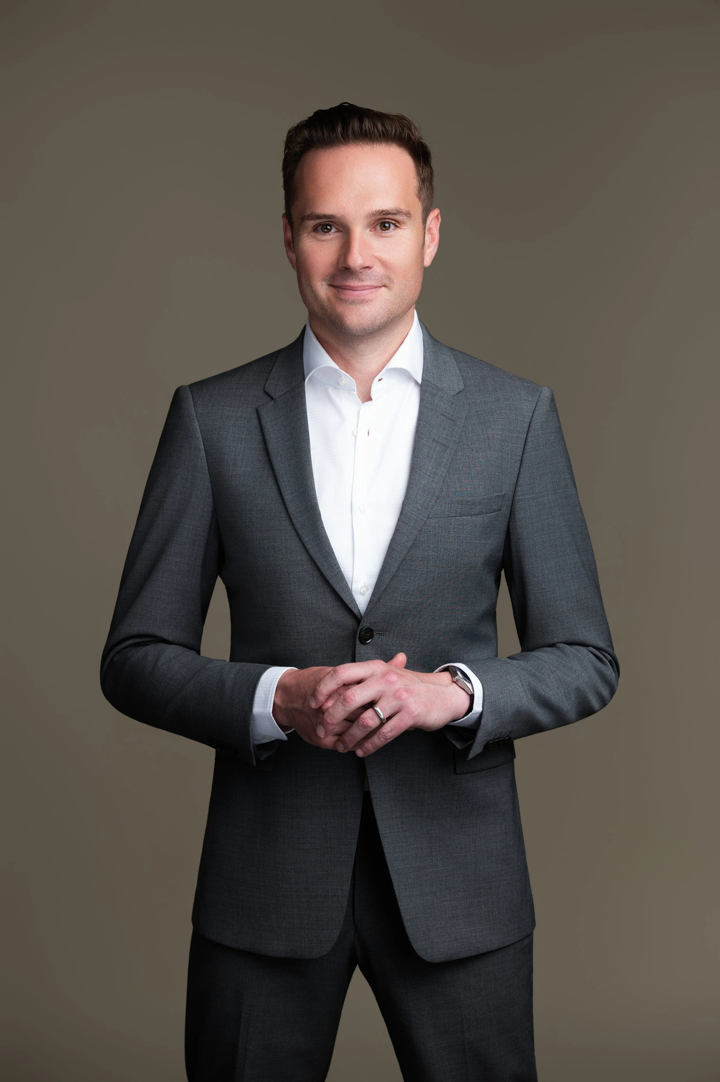 A professional man in a gray suit and white shirt, standing against a plain background, smiling softly with hands clasped.