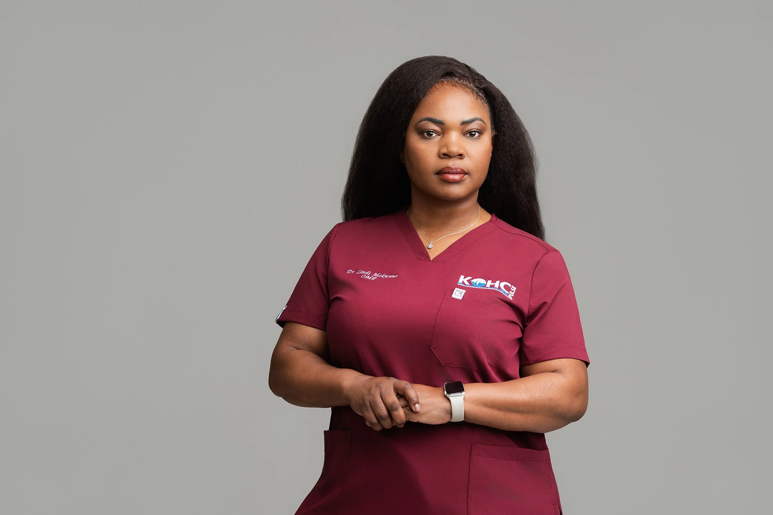 A woman dressed in medical scrubs with a maroon color, standing with arms crossed against a plain gray background, looking directly at the camera.