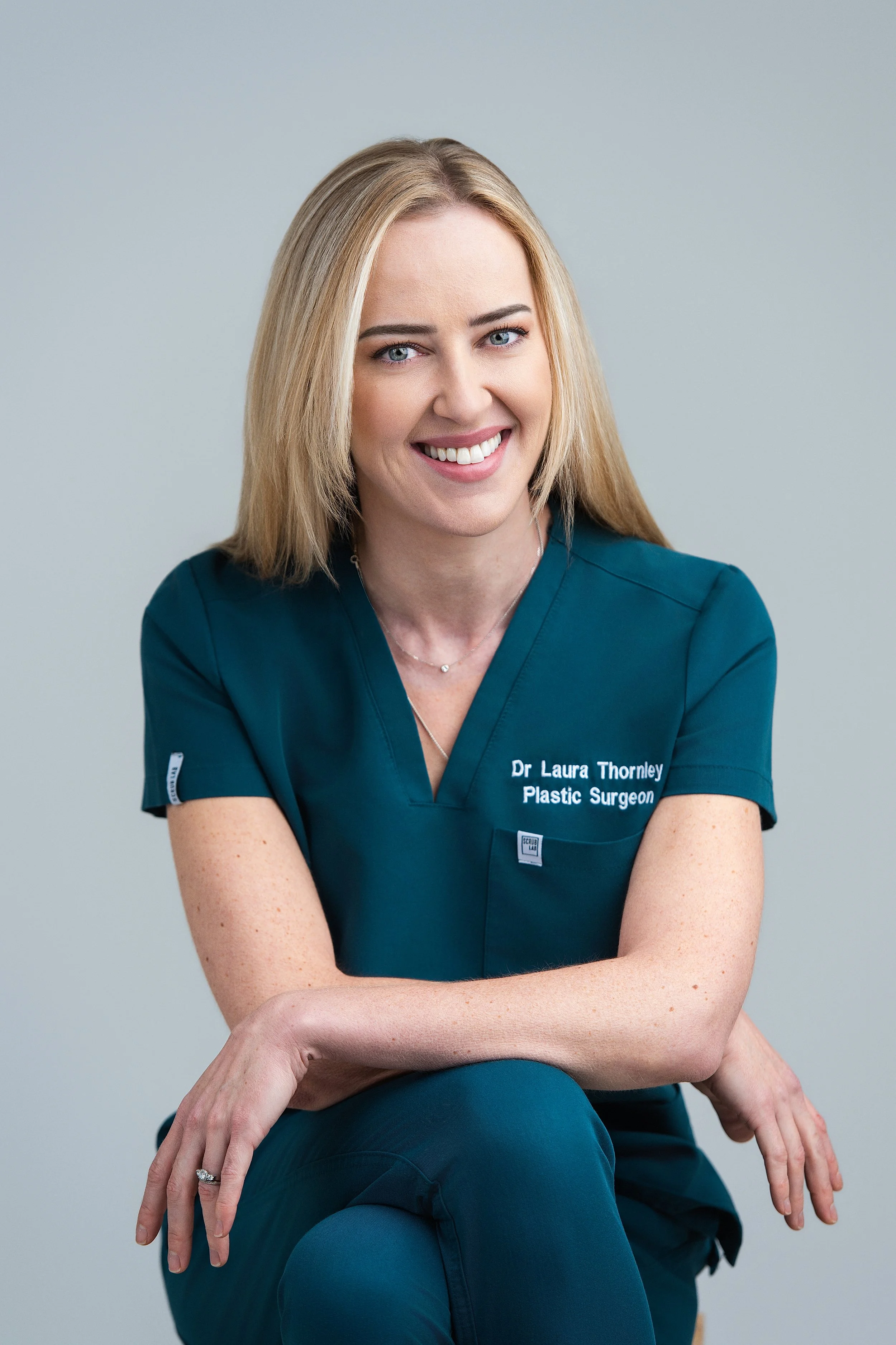 Photo of a woman with long blonde hair, wearing a dark teal medical scrub top with her name and title embroidered on it, smiling and sitting against a plain grey background.