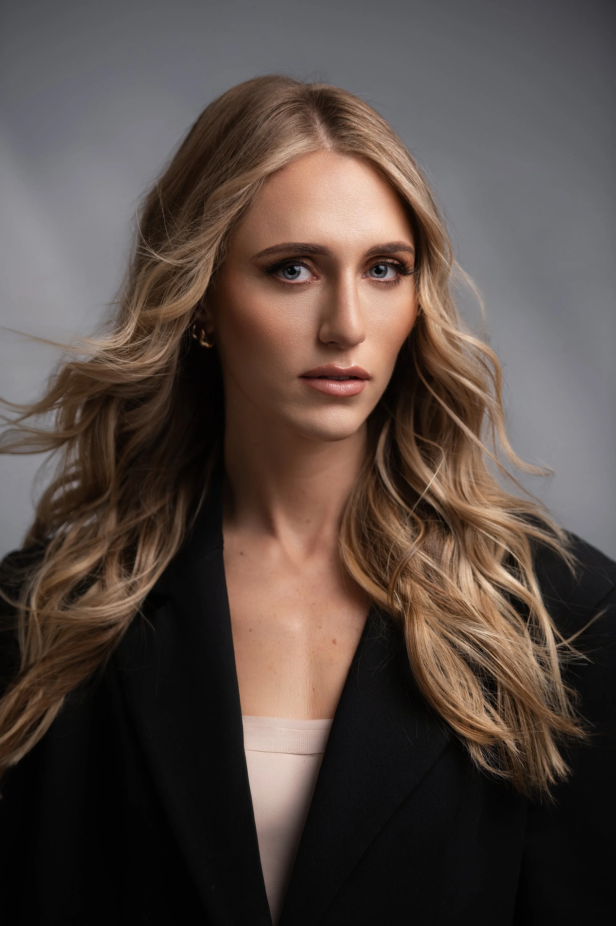 Close-up portrait of a woman with long, wavy blonde hair and blue eyes, wearing a black blazer over a cream top, against a gray background.