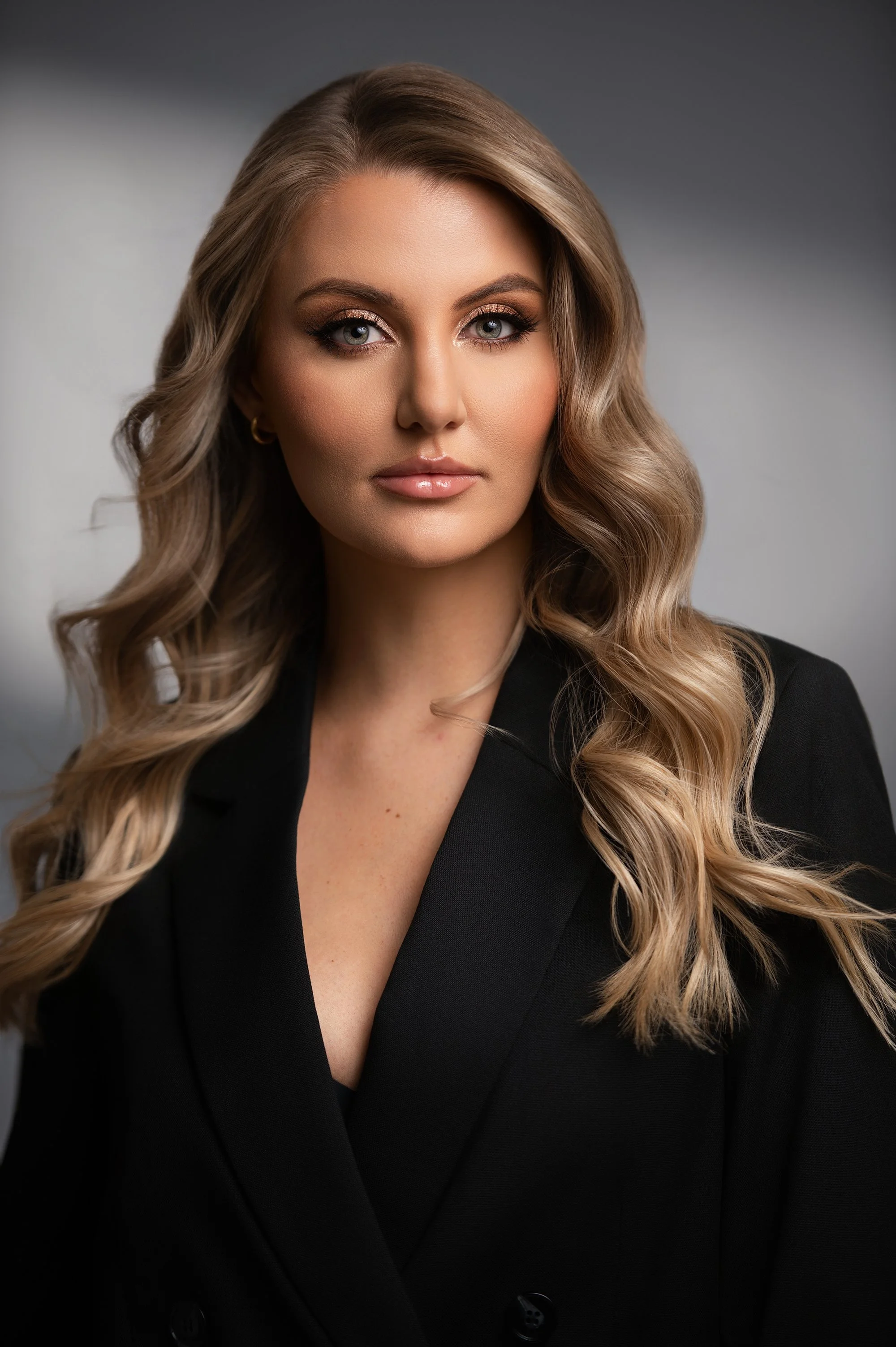 A woman with long wavy blonde hair, wearing a black blazer, and gold hoop earrings, posing in front of a gradient gray background.