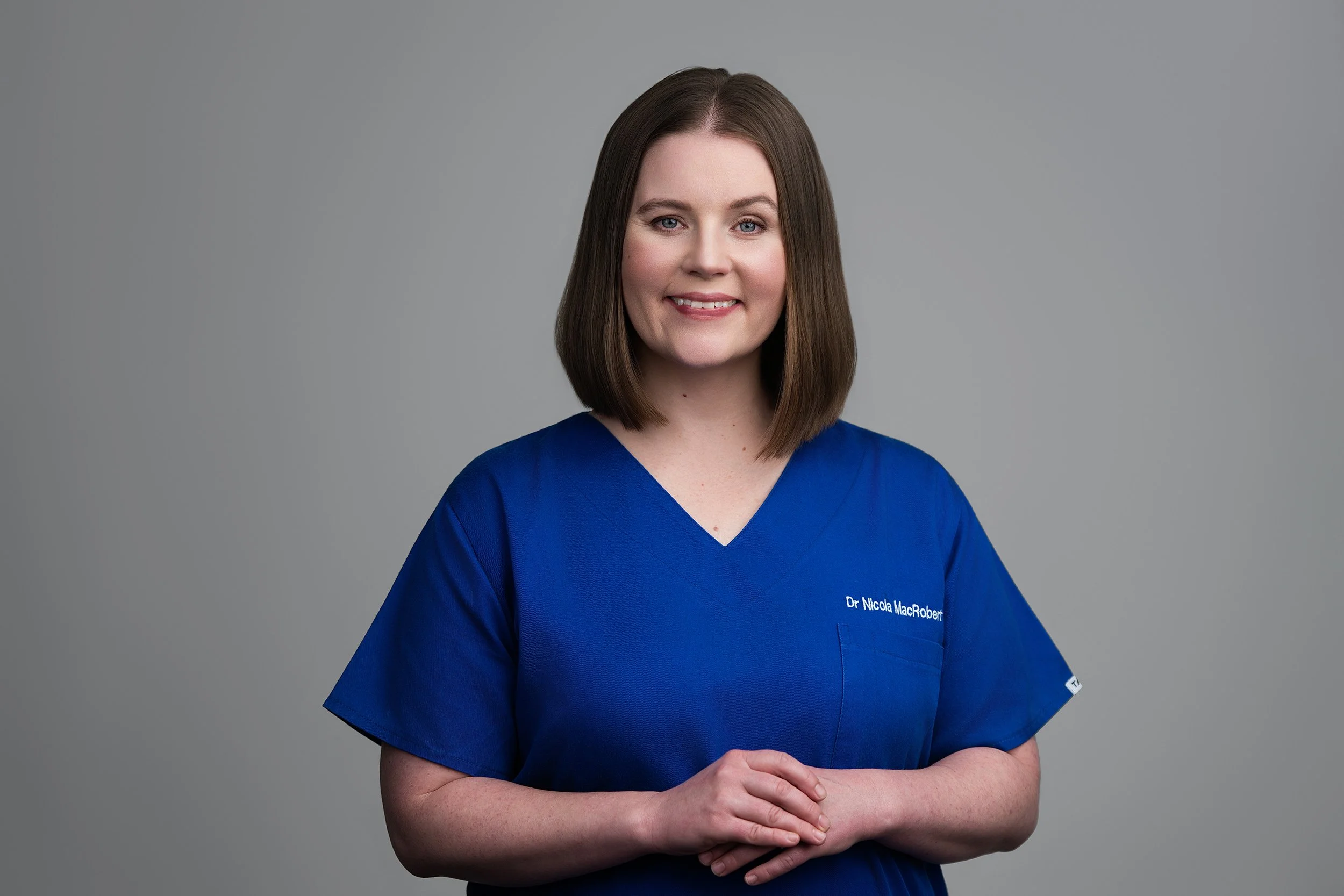 A woman with brown hair, wearing blue medical scrubs, standing in front of a gray background, smiling at the camera.