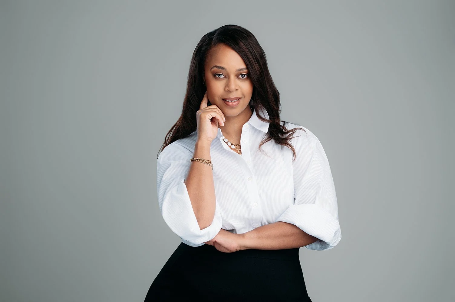 A woman with dark hair, wearing a white blouse and black skirt, posing with her hand near her face against a plain gray background.