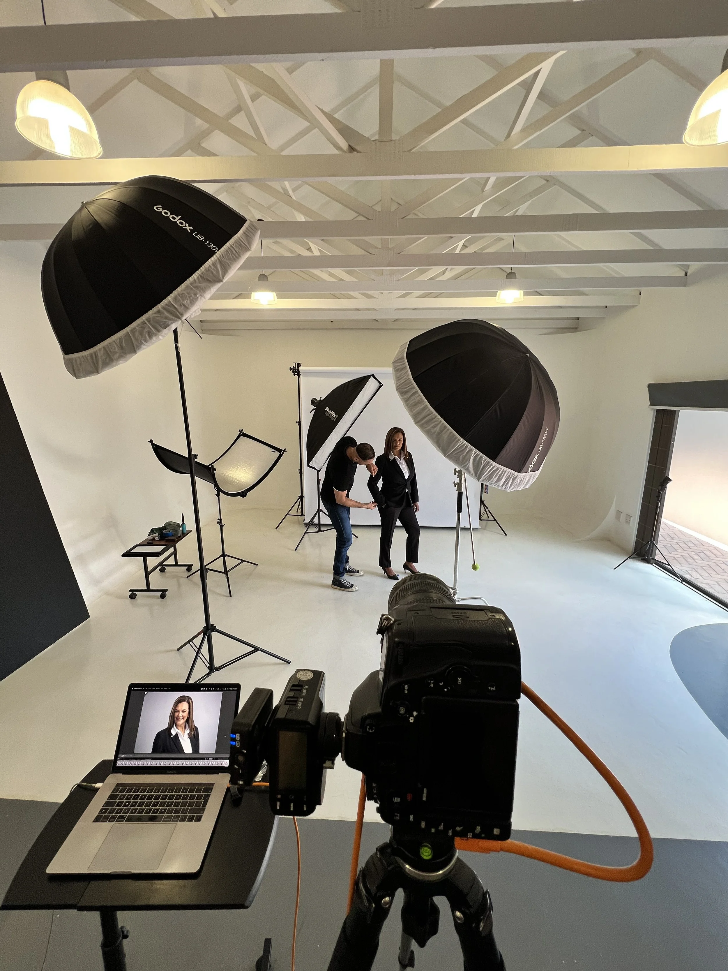 A photography studio setup with professional lighting equipment. A woman in professional attire is being photographed, standing in front of a white backdrop. There is a cameraman adjusting the woman’s pose. A camera on a tripod is focused on the woman, and a laptop displays her portrait in real-time. The room has a high ceiling with exposed white beams and multiple studio lights.