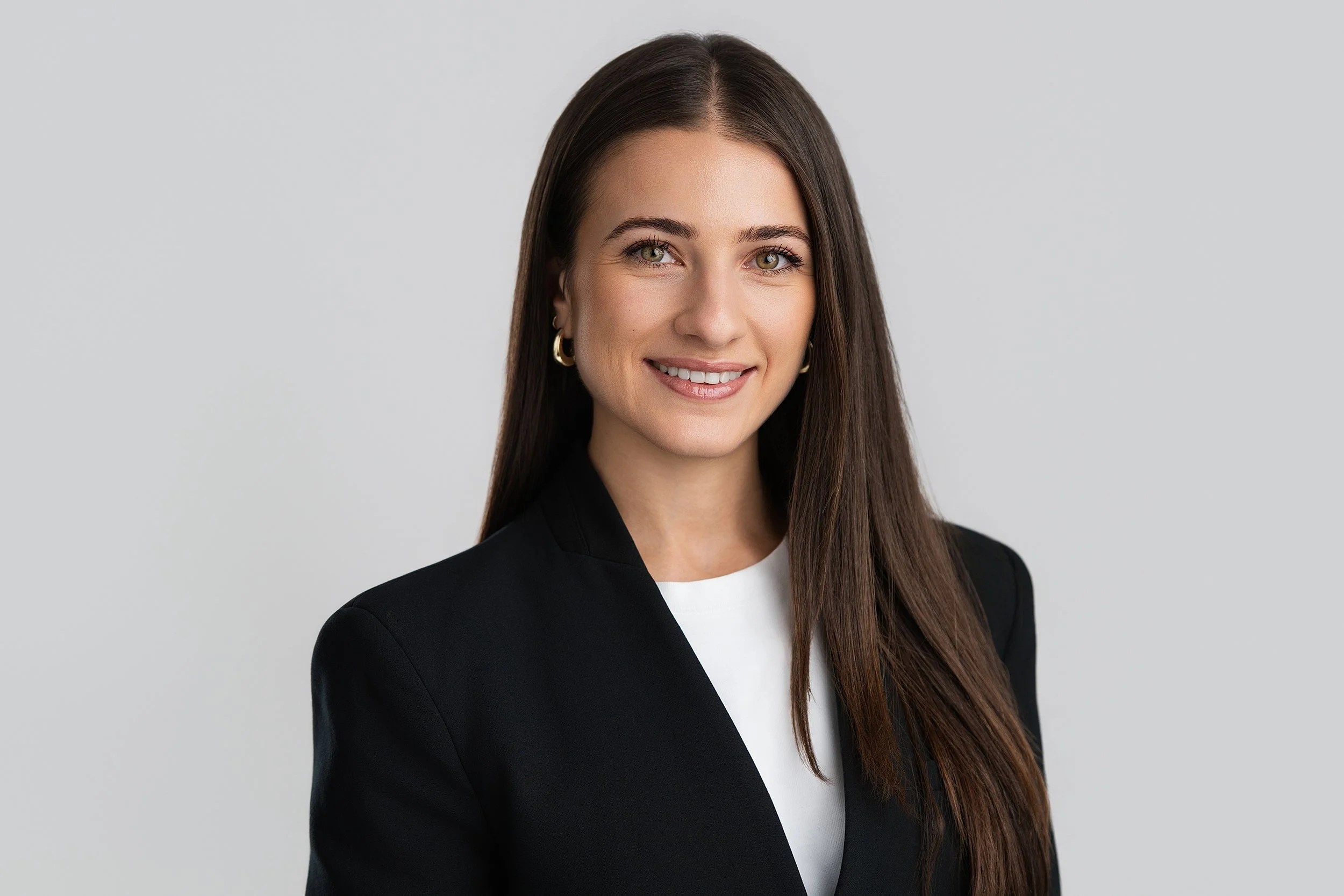 Professional woman with long brown hair, wearing a black blazer and white blouse, smiling against a light gray background.