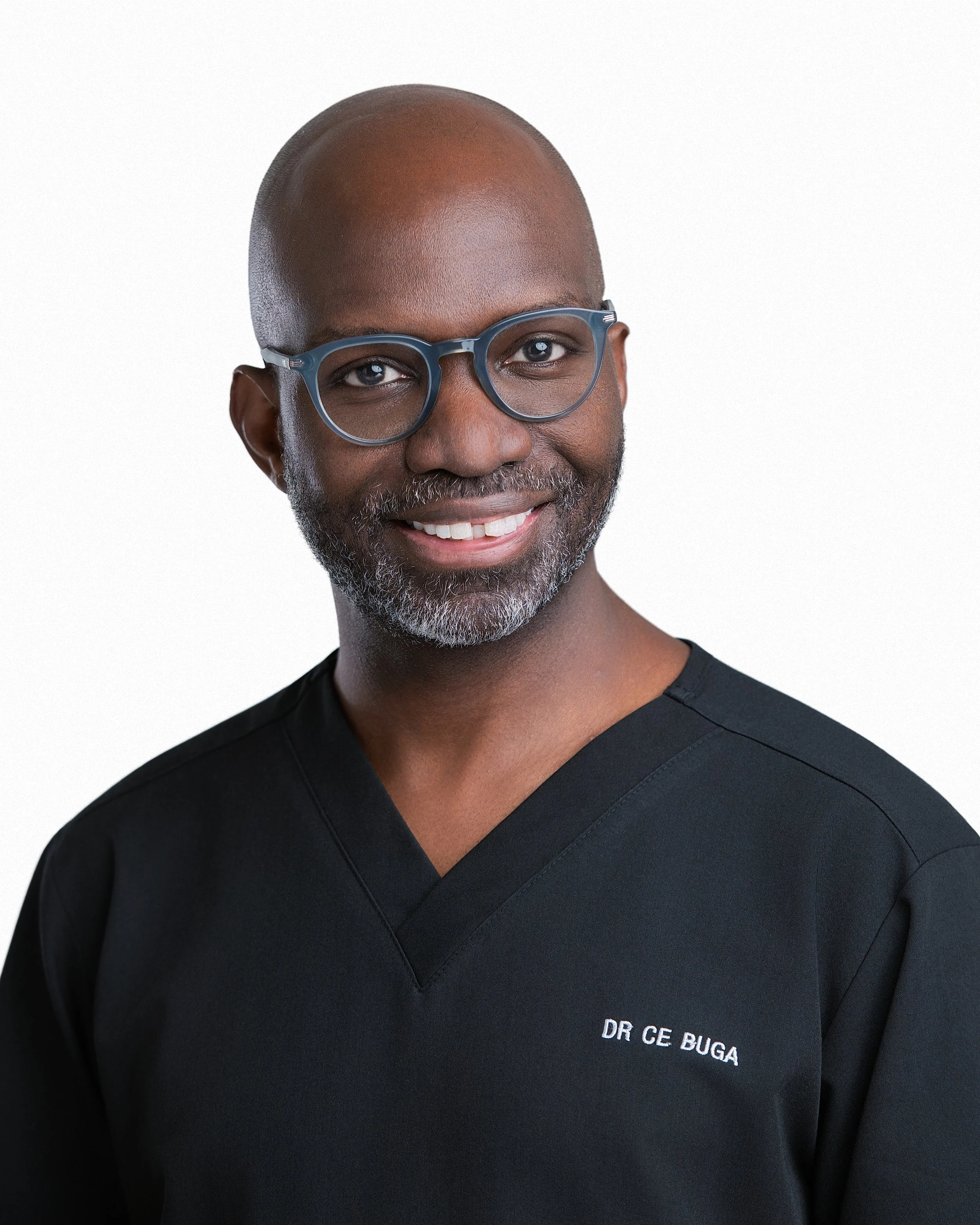 Close-up of an African-American man with a bald head and glasses, smiling, wearing a black medical scrub with the embroidered name 'DR CE BUGA'.