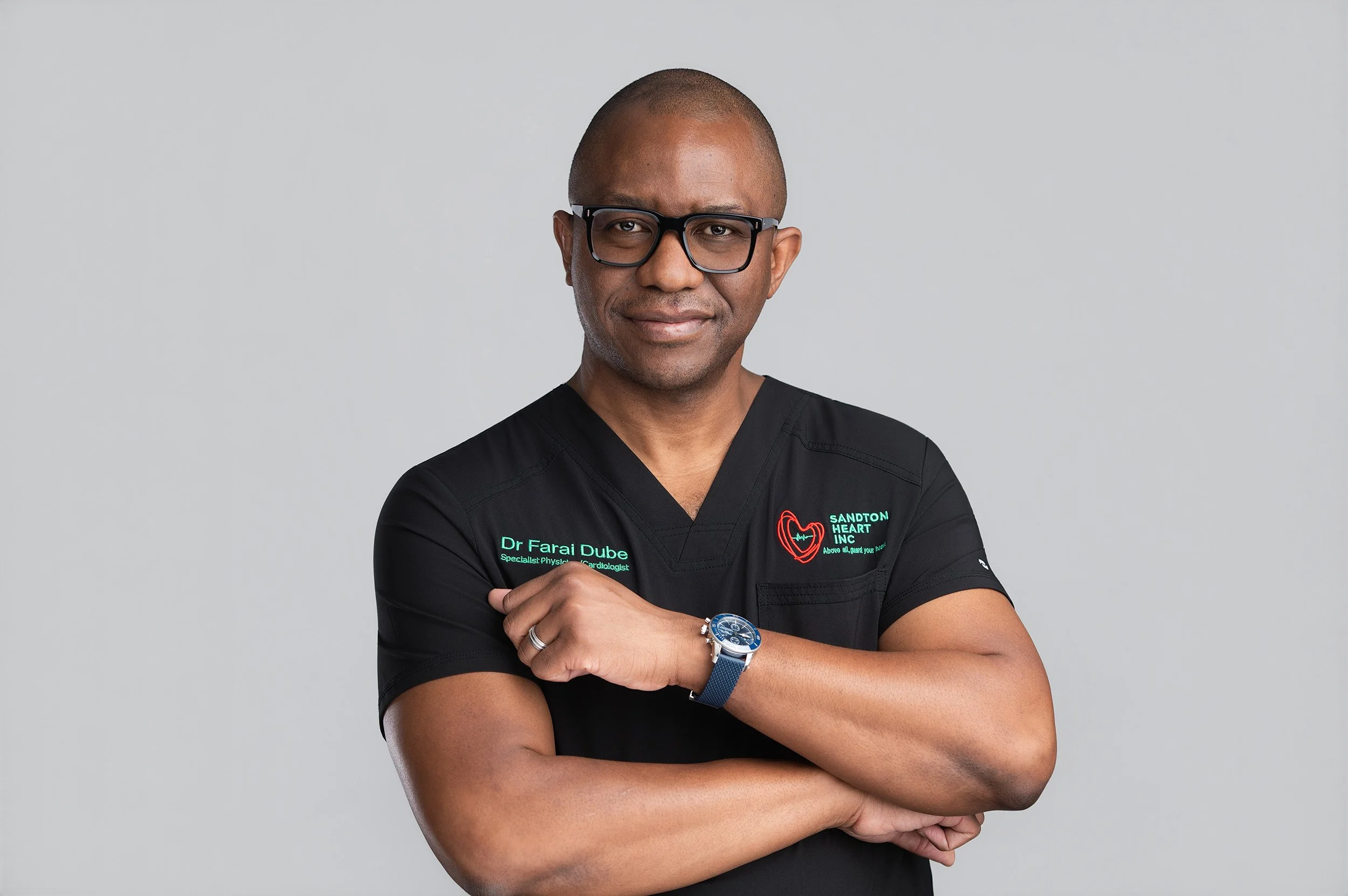 A man with glasses and a shaved head wearing black medical scrubs with green text that reads 'Dr. Farai Dube, Specialist Physiologist, Cardiologist' and a logo with a heart, standing against a plain light gray background. He has his arms crossed and 