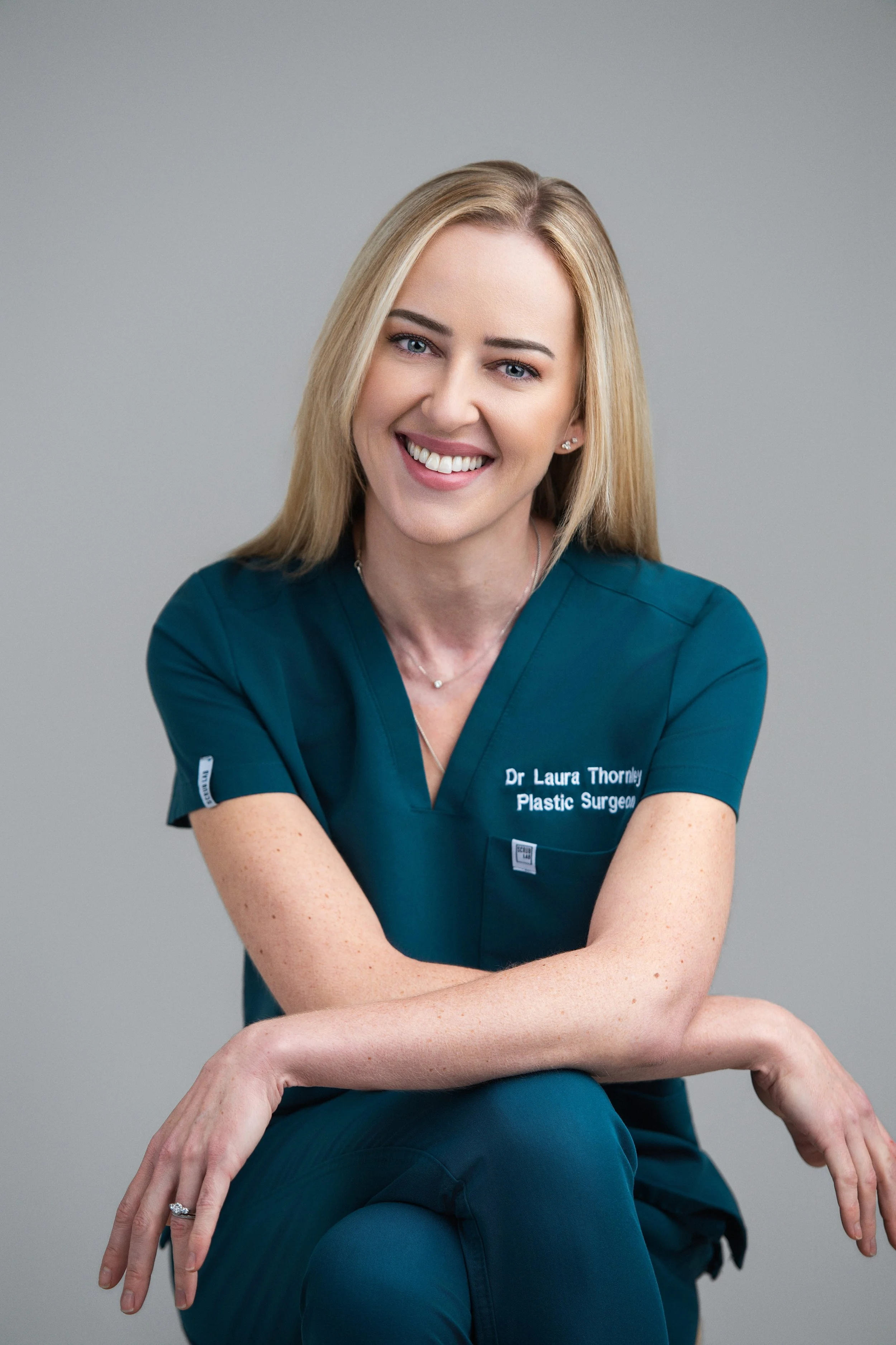 A smiling woman with blonde hair, wearing a dark teal medical scrub top, sitting against a neutral gray background. She has a name badge with "Dr Laura Thornley, Plastic Surgeon" on it.