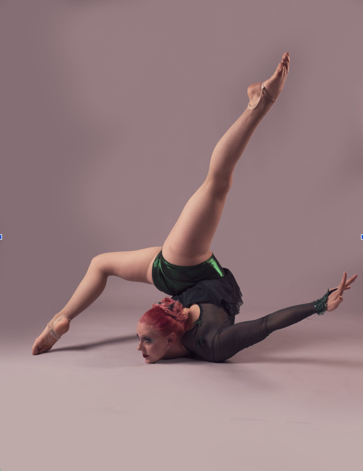 A female gymnast with pink hair performing a contortion pose with one leg extended upwards and head resting on the ground, against a plain pink background.
