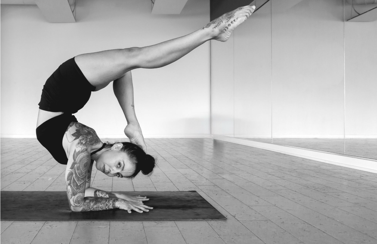 A woman with tattoos practicing yoga in a studio, balancing on her forearms with one leg extended upward and the other bent, demonstrating advanced yoga pose.