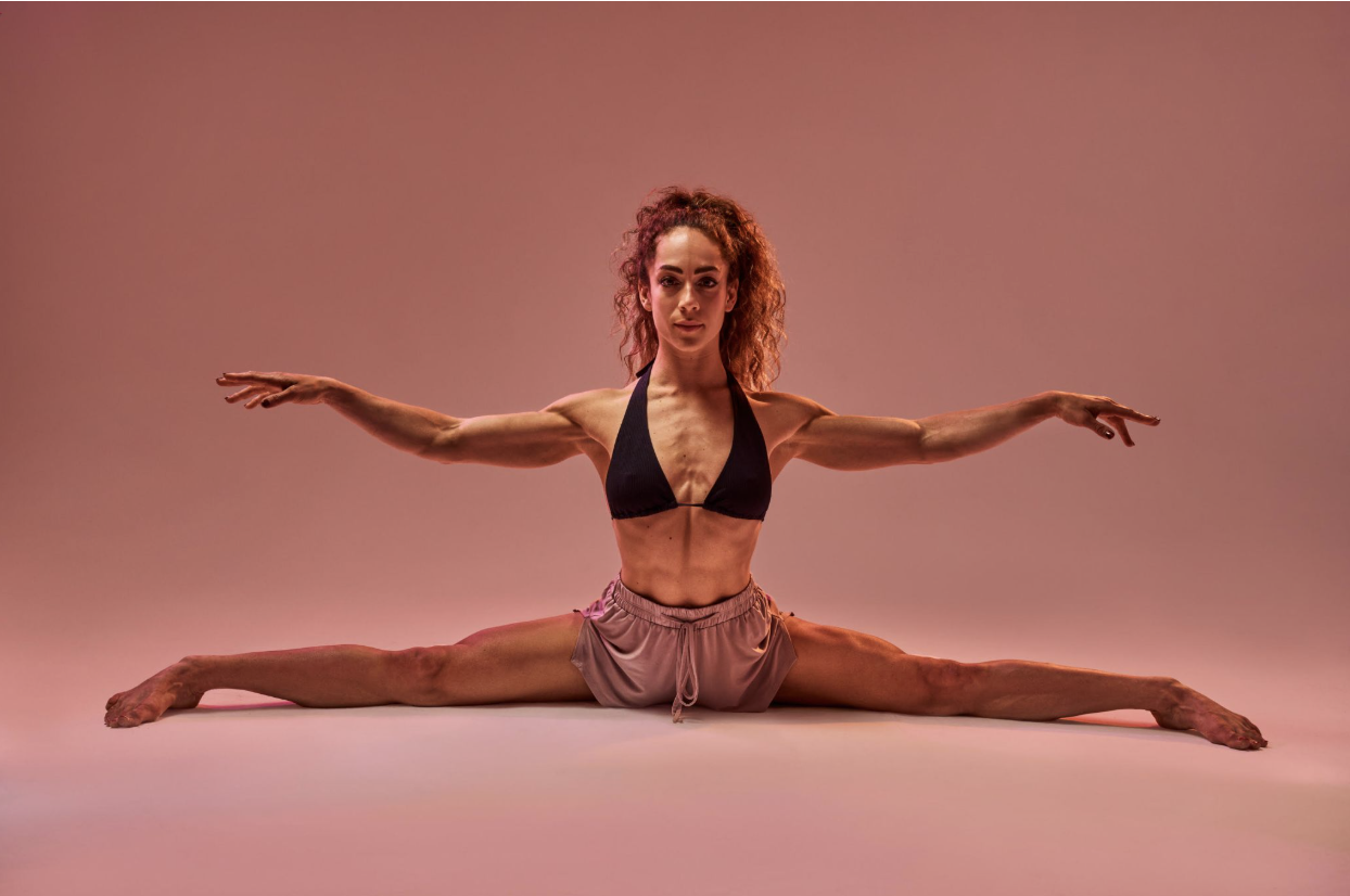 A woman with curly hair doing a full split and a stretch with arms extended sideways, wearing a black bikini top and pink shorts, against a pinkish background.
