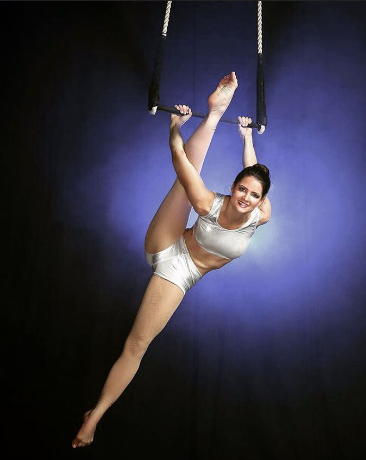 A woman in shiny athletic wear, hanging upside down on a trapeze, smiling, against a blue background.