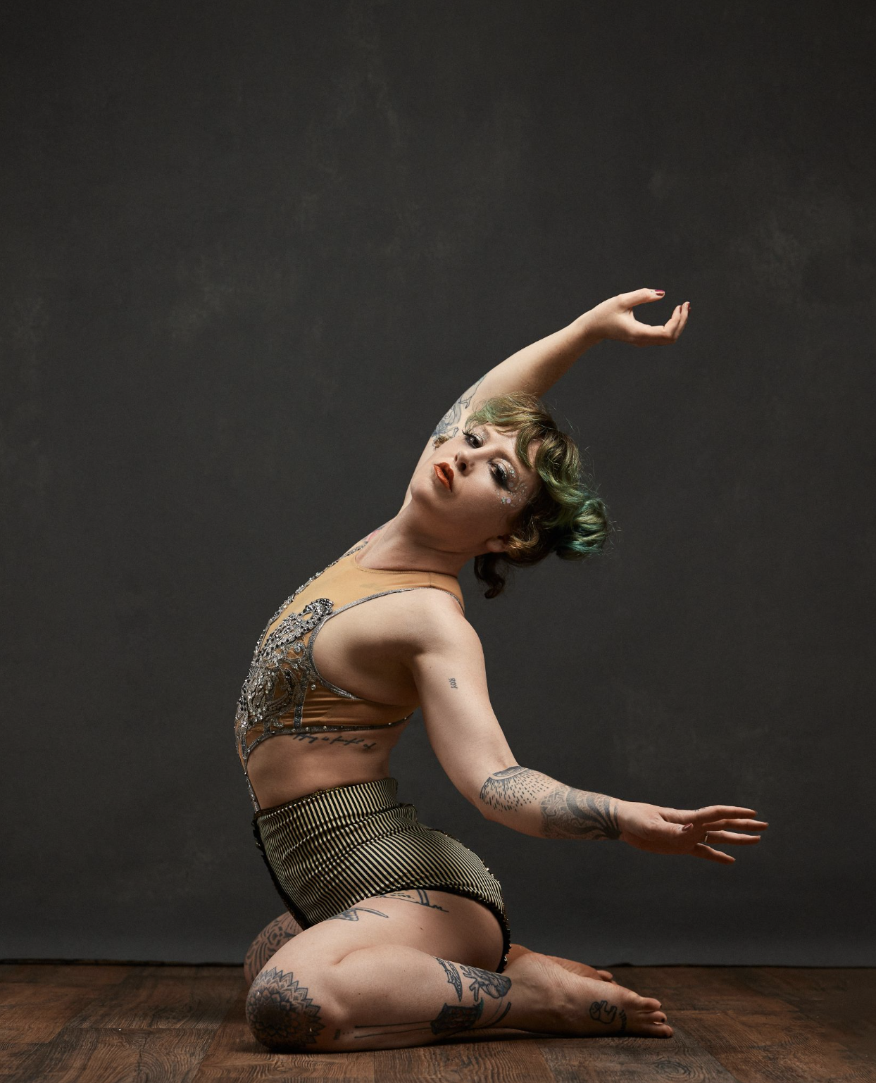 A woman with tattoos in a dancer pose, kneeling with one arm raised and the other extended, against a dark background.